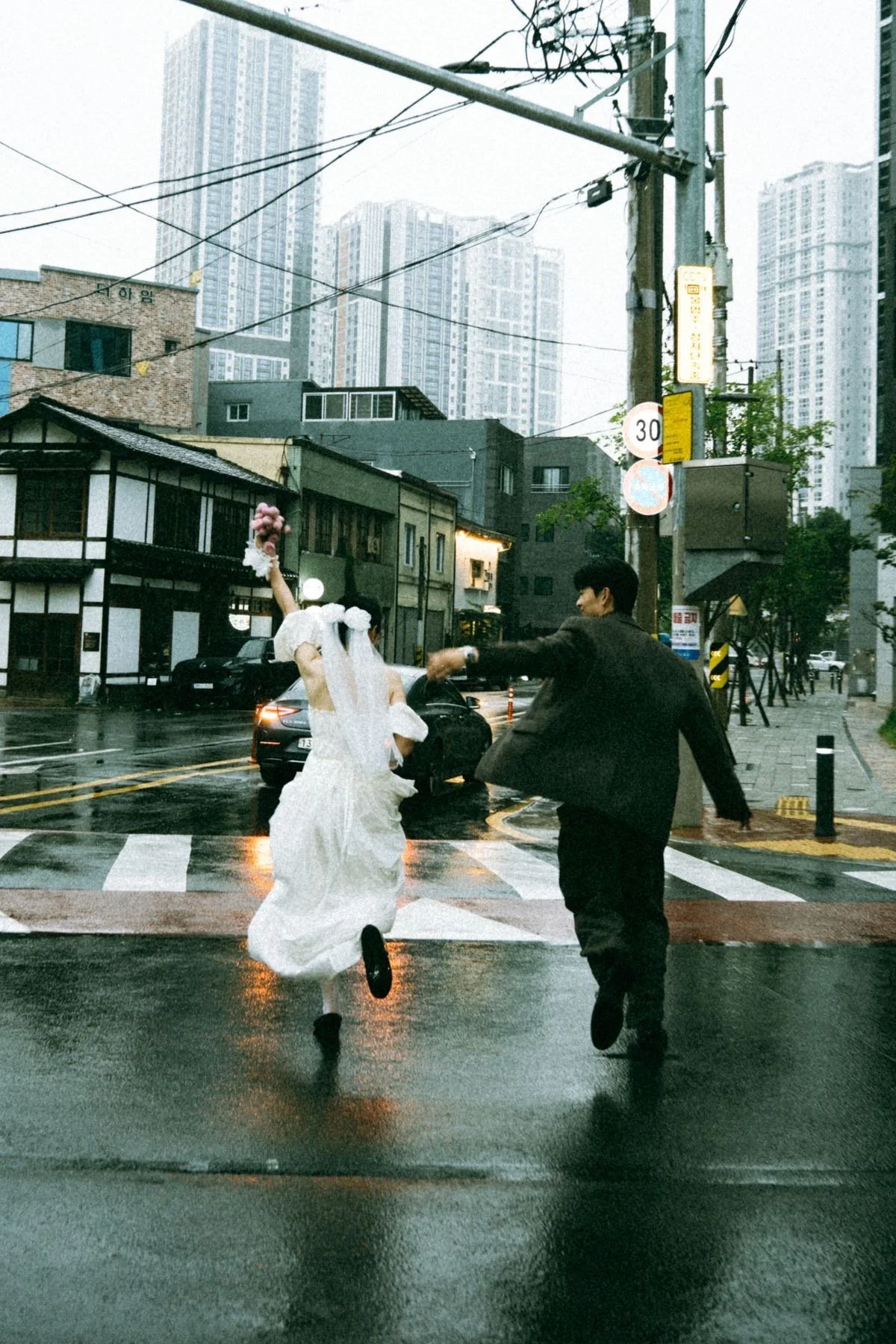 A bride and groom run across a wet crosswalk on a rainy city street, smiling with buildings and trees in the background.