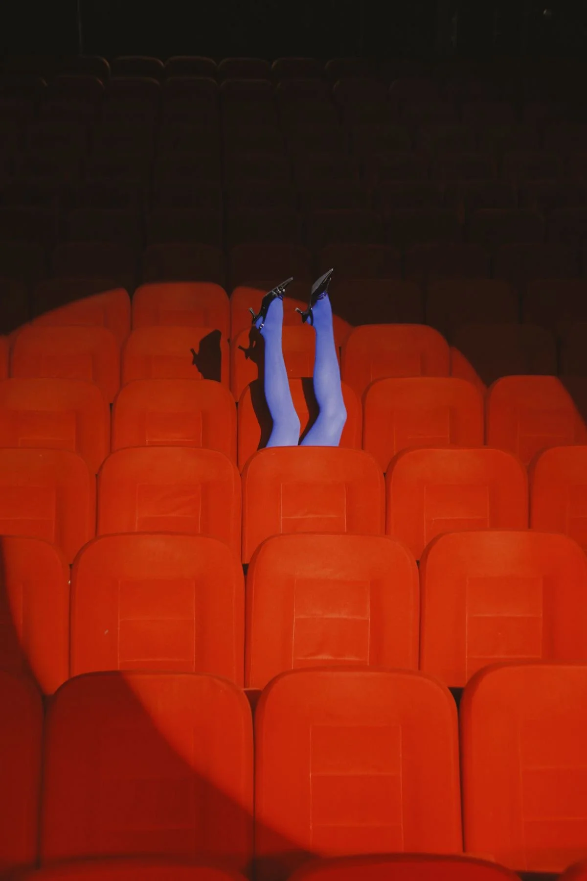 Person lying on red theater seats with legs raised, wearing blue stockings and black high heels, in an empty theater.