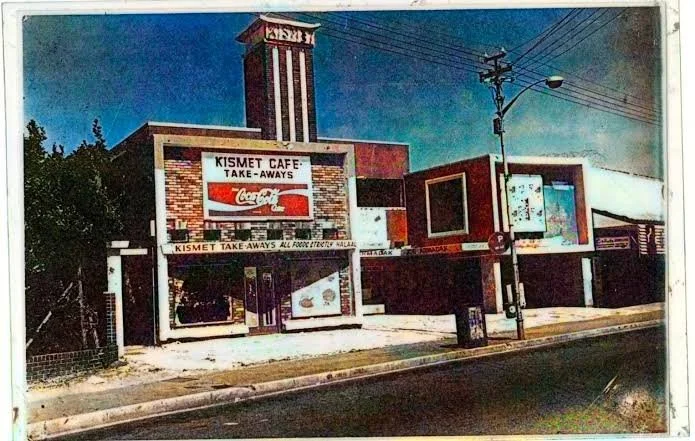 A vintage photo of Kismet Cafe, a small restaurant with a brick facade, a Coca-Cola sign, and a drive-thru window, located on a city street with a visible clock tower in the background. The kismet cinema located in Athlone, Cape Town.