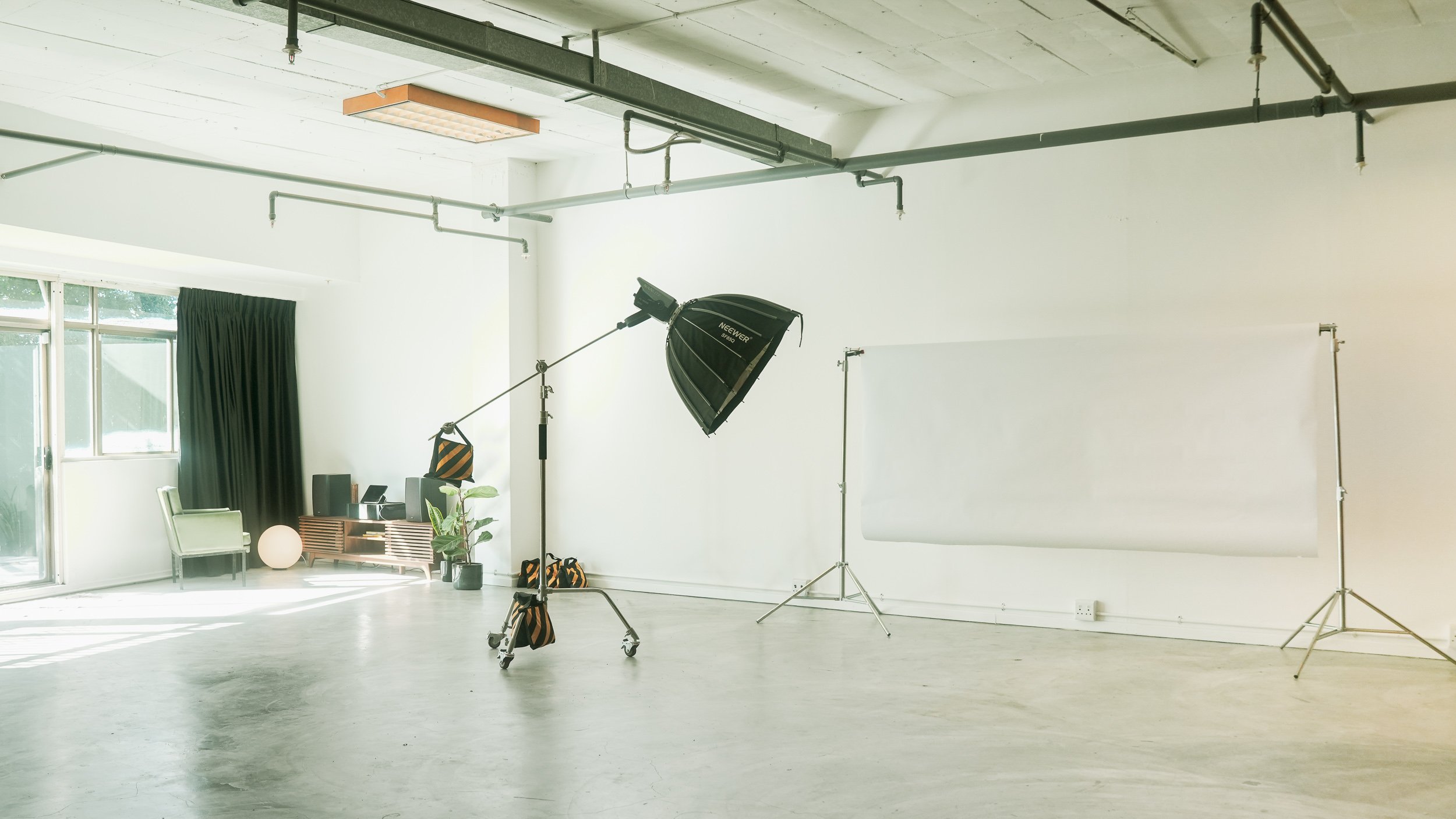 Photography studio with a large white backdrop, professional lighting equipment, and minimal furniture including chairs, a plant, and a spherical lamp near a window with dark curtains. Professional studio in Athlone, Cape Town