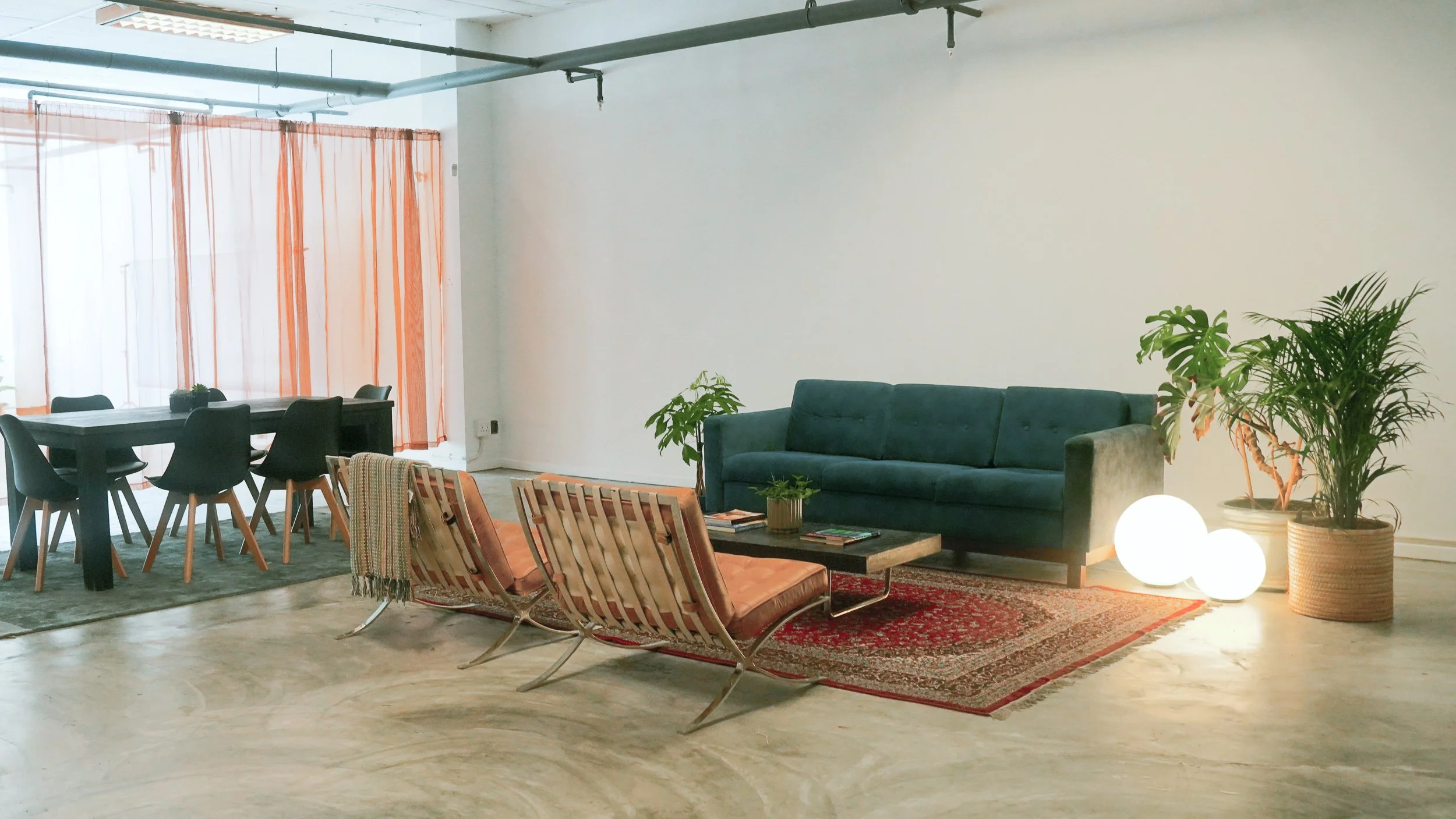 An interior living space with a dark blue sofa, wooden armchairs, a coffee table with books, indoor plants, floor lamps, and a window with orange curtains. Lounge in film studio. Production company based in Athlone South Africa