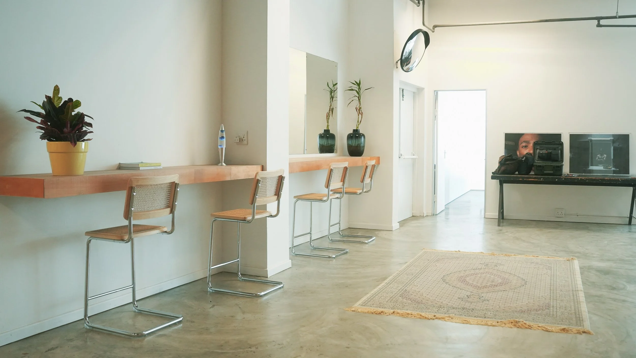 A minimalistic room with white walls and light wood flooring, featuring bar stools at a wooden counter with plants and a lava lamp, and a black table with camera equipment and a reflection of a person's face. Hair and Make-up counter in film studio