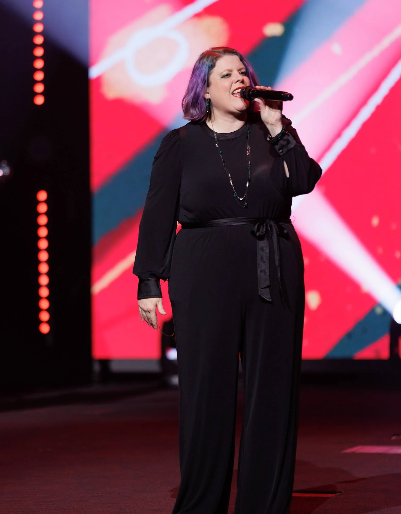 A woman with purple hair singing into a microphone on stage, with a colorful abstract background.