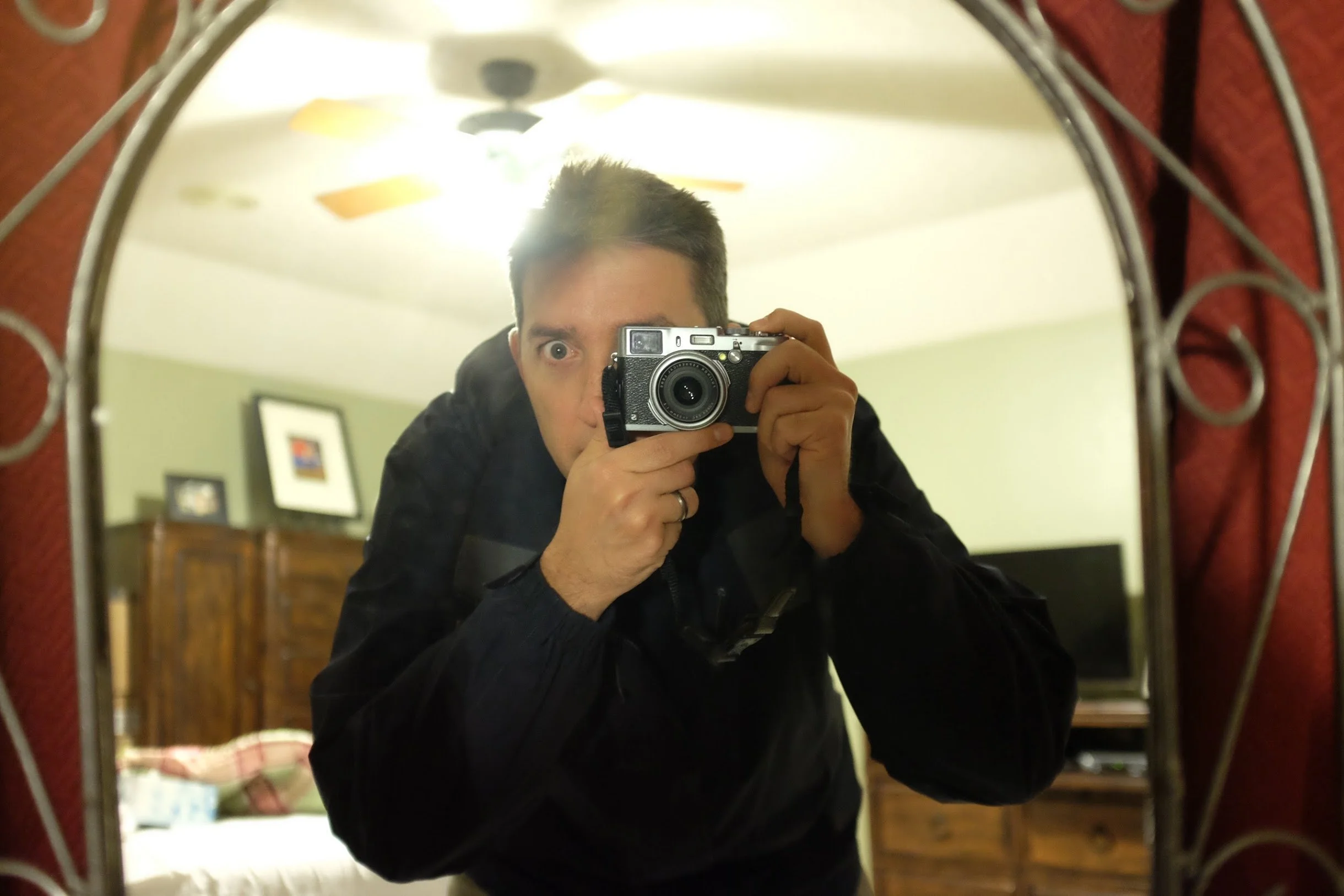 Person taking a photograph of themselves in a mirror using a camera, reflecting a room with a ceiling fan and framed pictures.