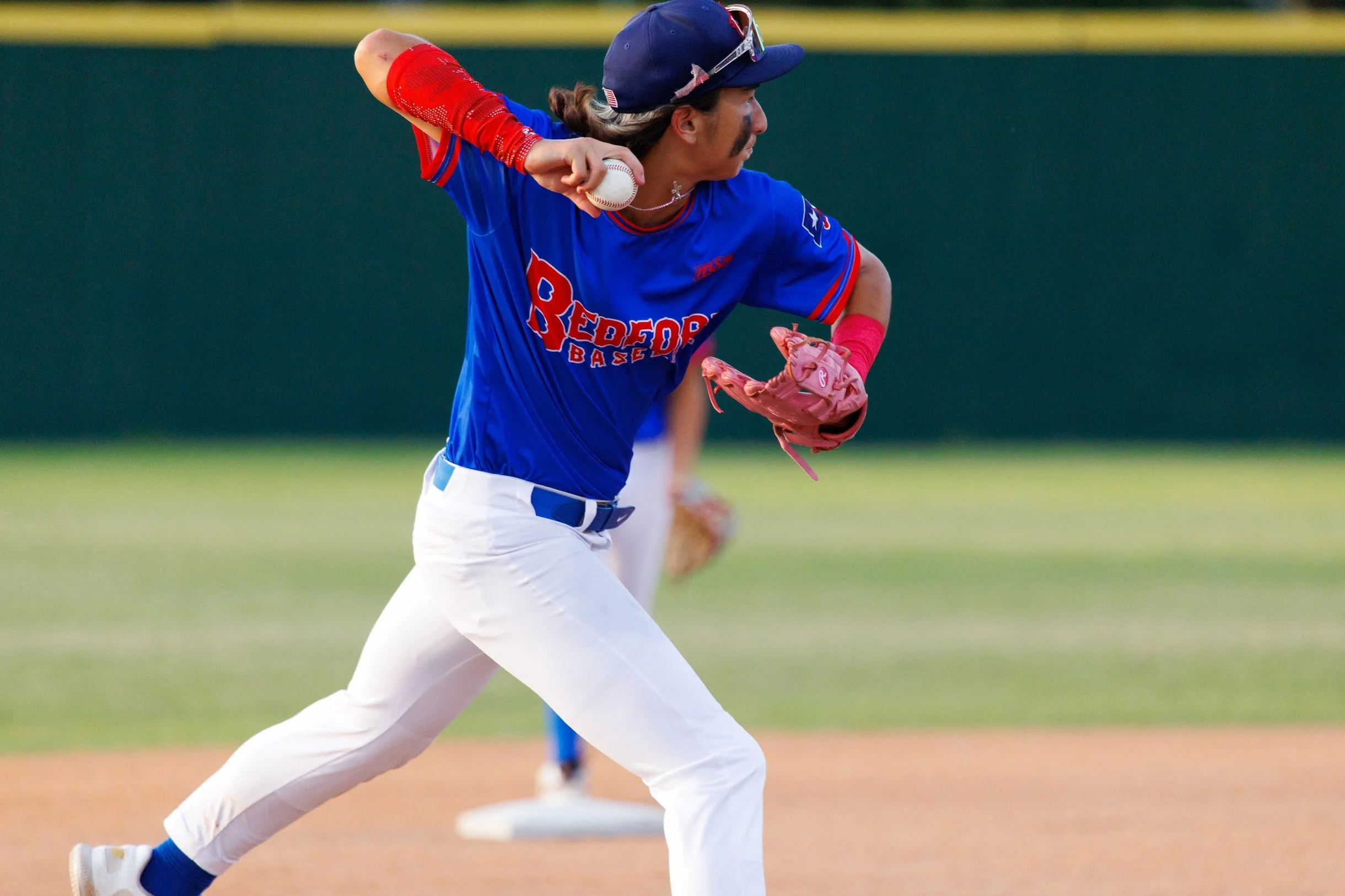 A baseball player in a blue and white uniform running on a baseball field with a baseball in hand, wearing pink gloves and a baseball cap with sunglasses on top.