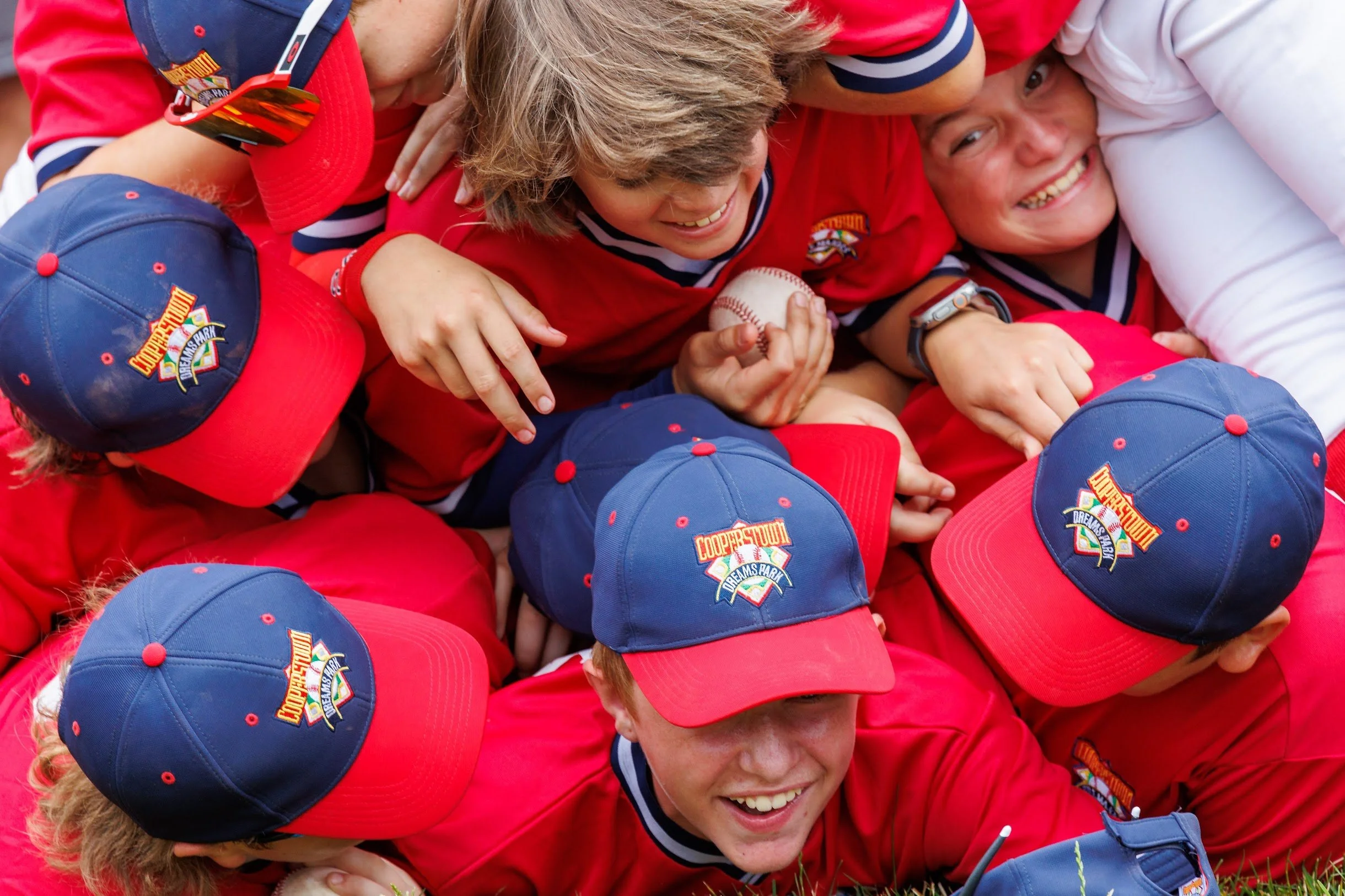 Children wearing red and blue baseball uniforms and hats, participating in a friendly rugby game on a grassy field.