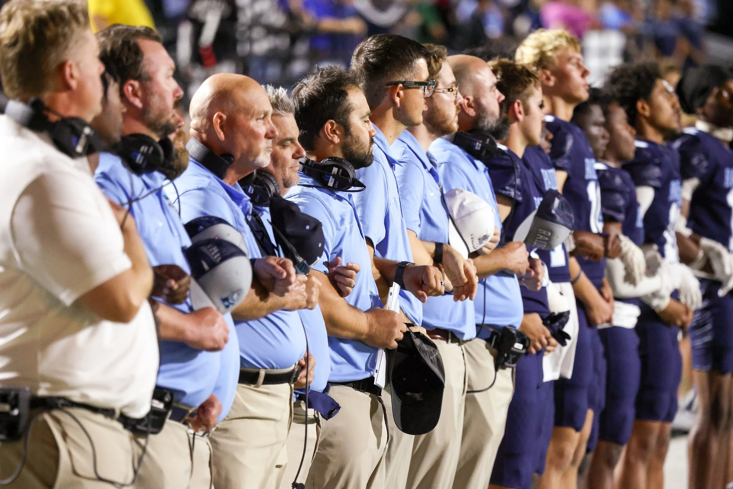 A row of football coaches and team staff members standing during a game, all wearing matching light blue and navy uniforms, some holding helmets and wearing headsets.