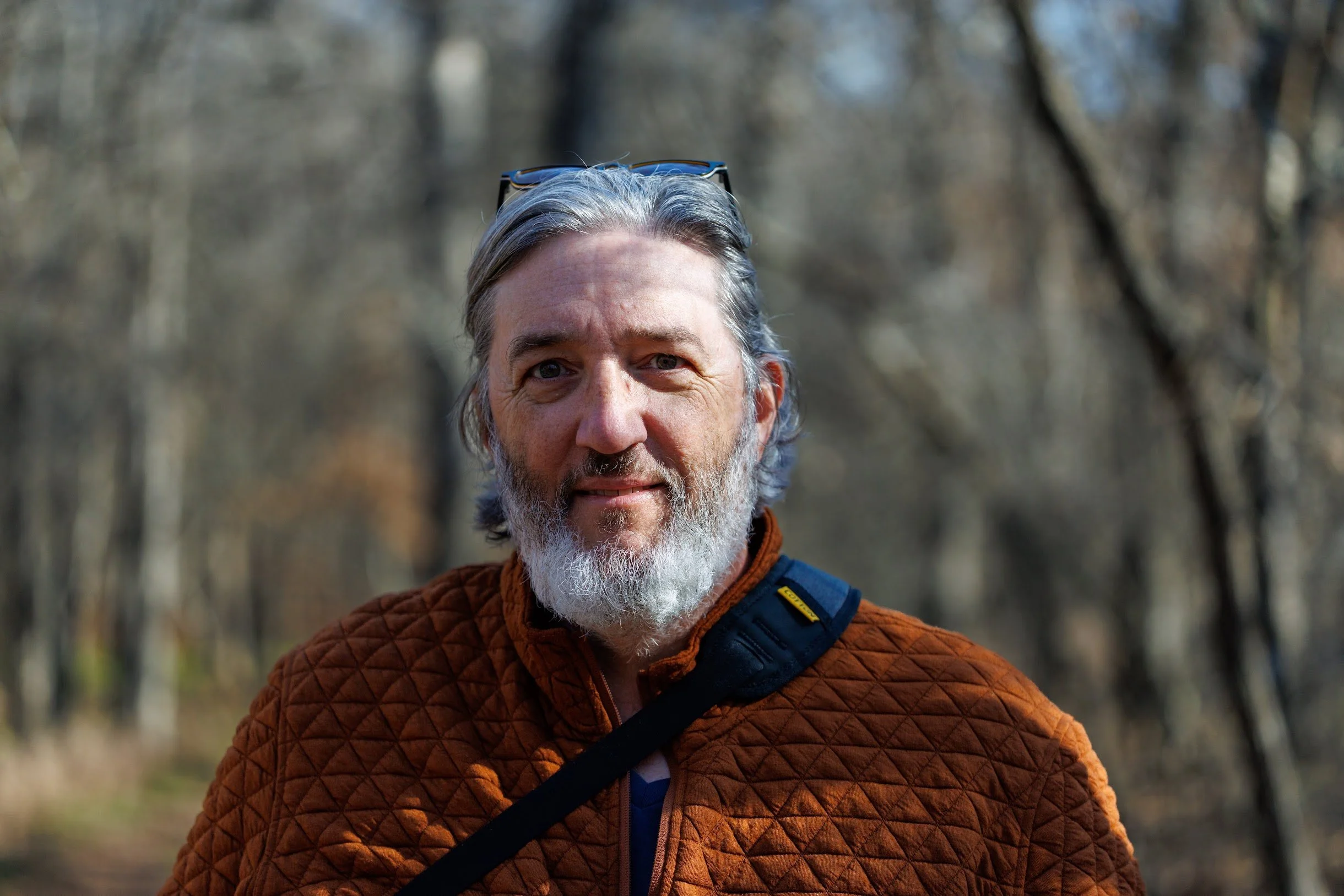 A man with gray hair and a beard wearing a brown quilted jacket and glasses on his head, outdoors in a wooded area during daytime.