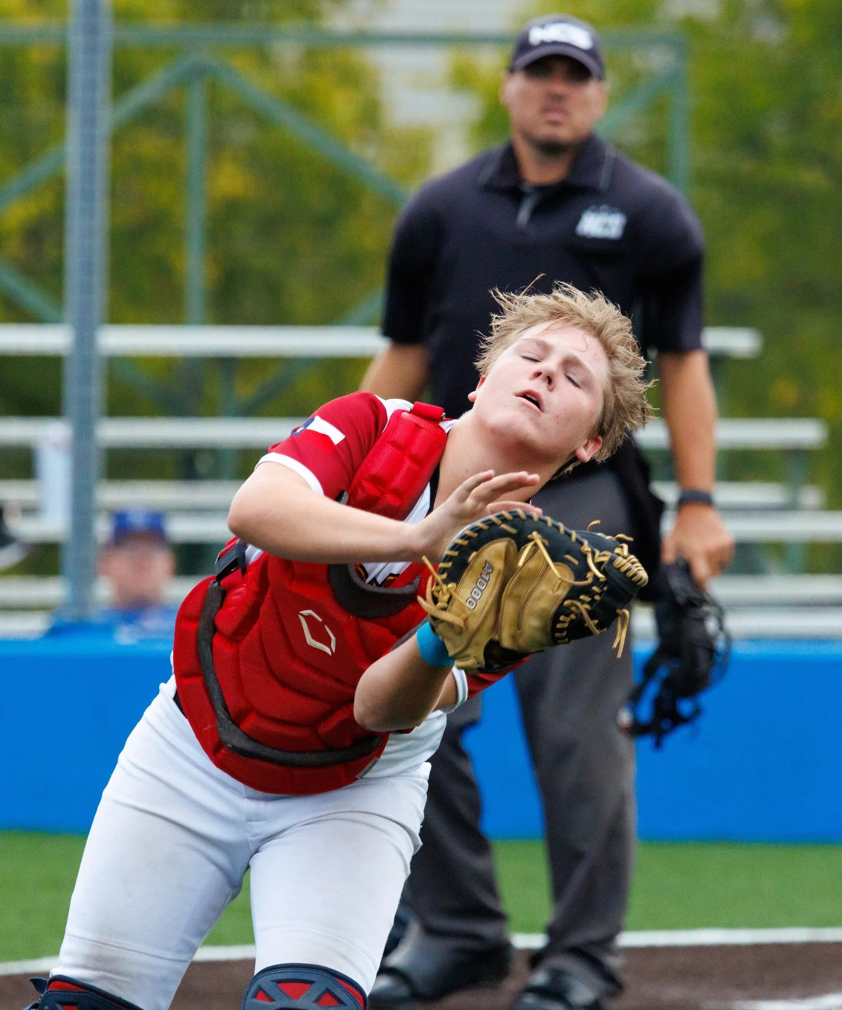 A young baseball player in a red and white uniform is crouching with his glove, focused on the game, while an umpire stands behind him on a green baseball field.