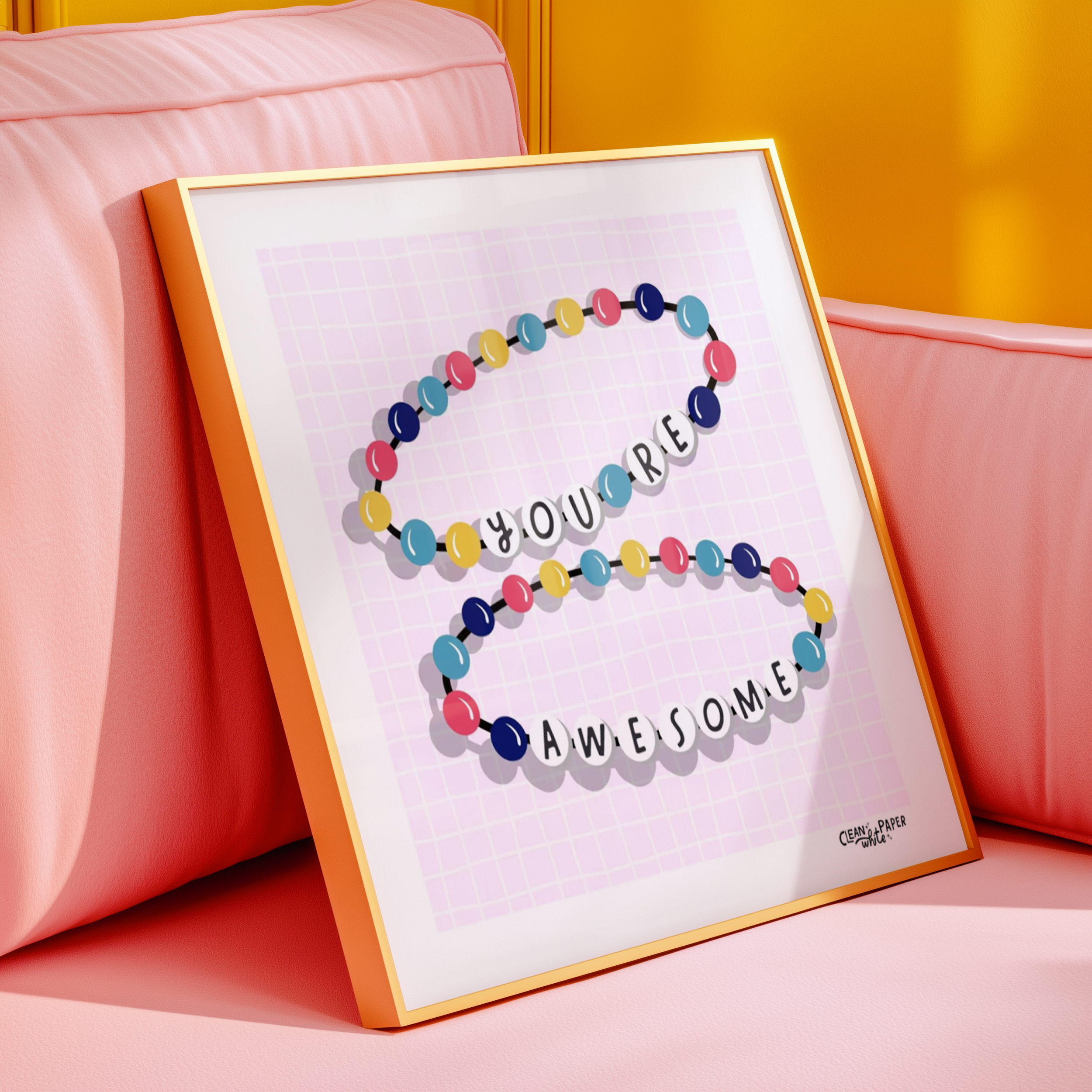 Colorful framed art piece with a circular string of beads spelling out 'YOU'RE AMAZING' on a pink grid background, leaning against a pink upholstered headboard with a yellow wall behind it.