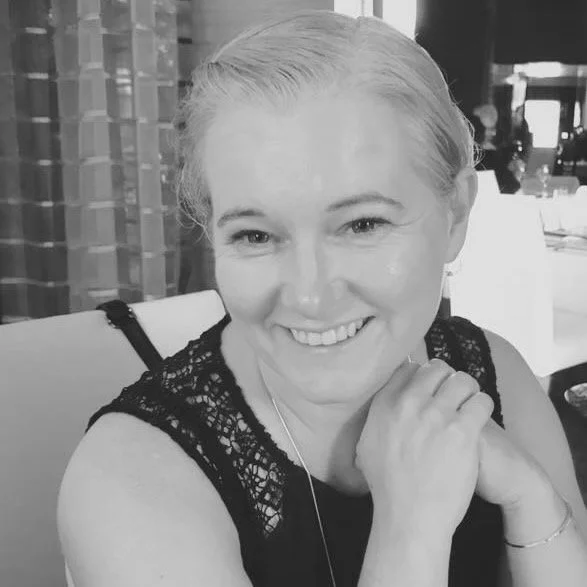 A woman with braided blonde hair smiling and resting her chin on her hand, in a black lace top, in a restaurant setting.