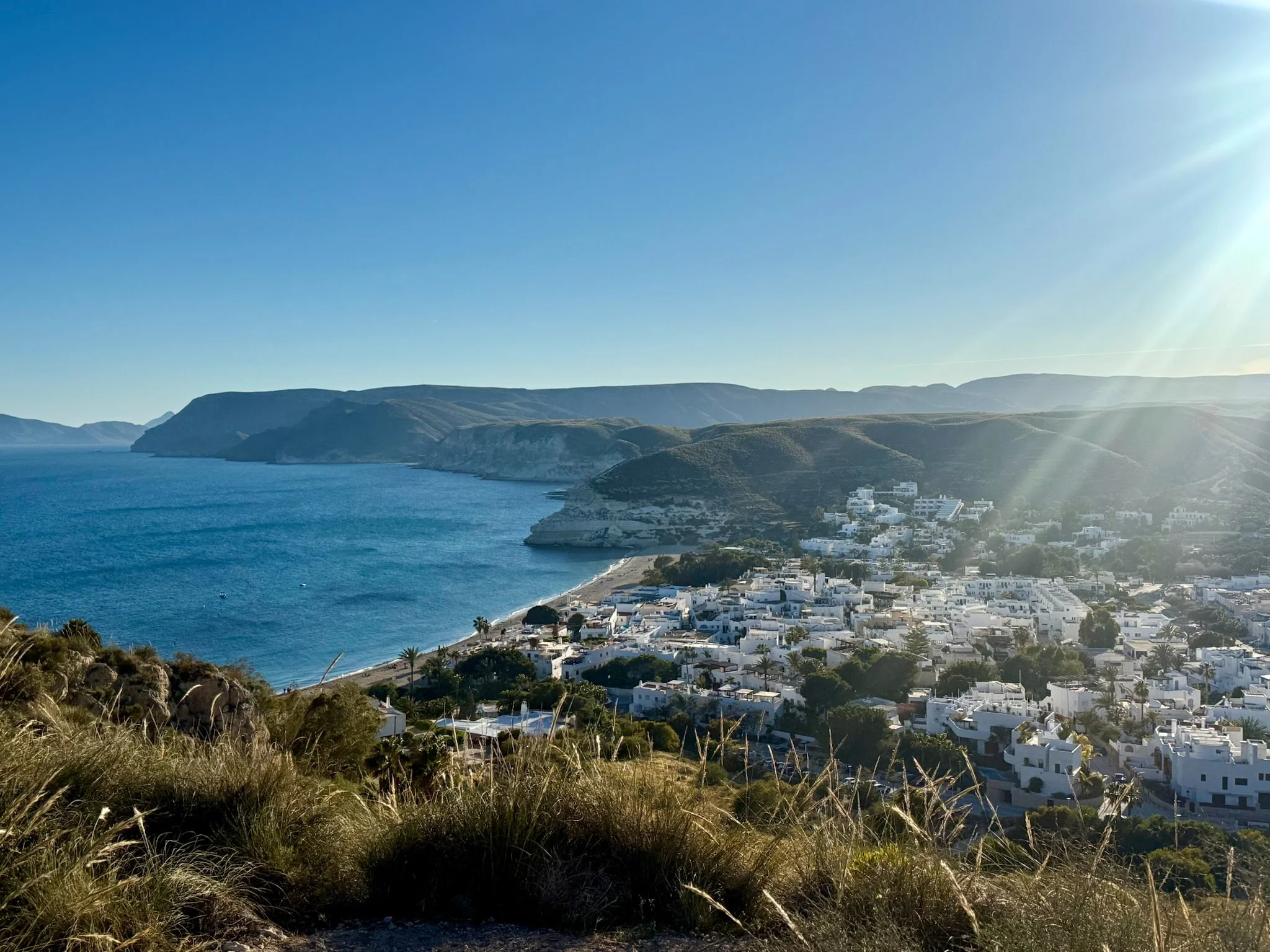 Coastal landscape with blue sea, hilly terrain, white buildings, and clear sky with sunlight