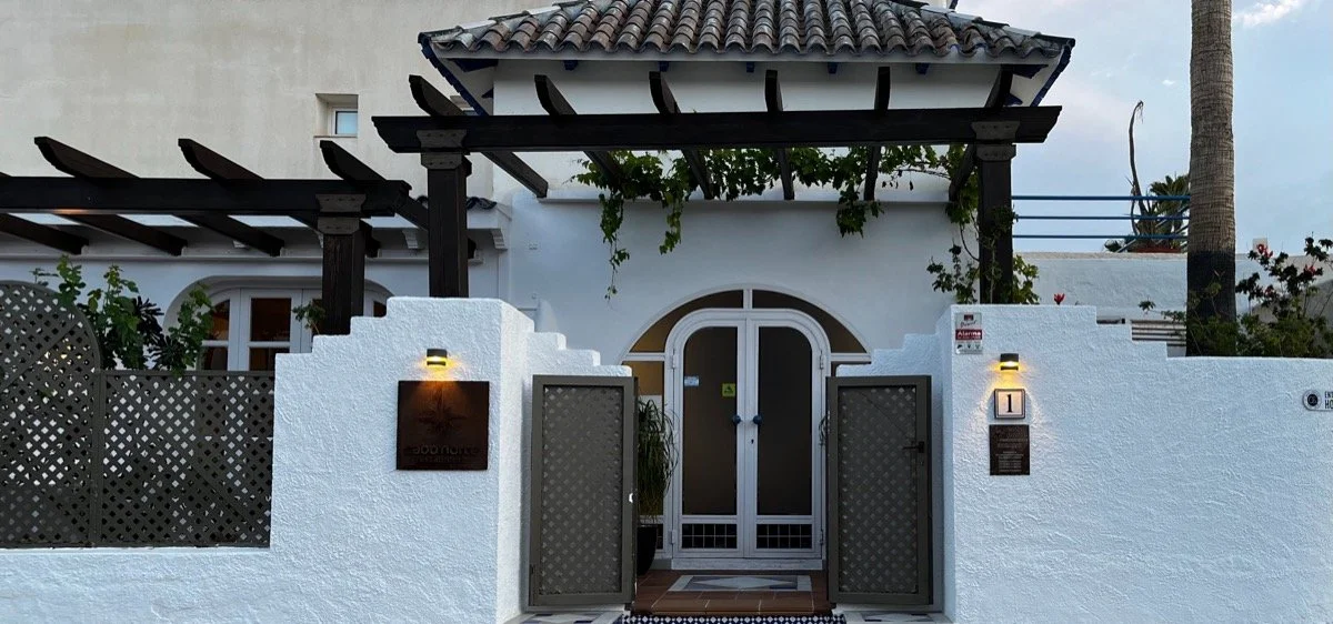 White stucco house with arched white door, black metal gate, outdoor lights, wooden pergola with green climbing plants, palm trees, and flowering plants in the background.
