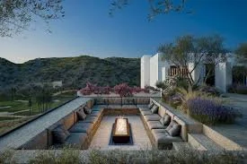 Outdoor patio with built-in seating around a fire pit, surrounded by landscaped gardens and hills in the background.