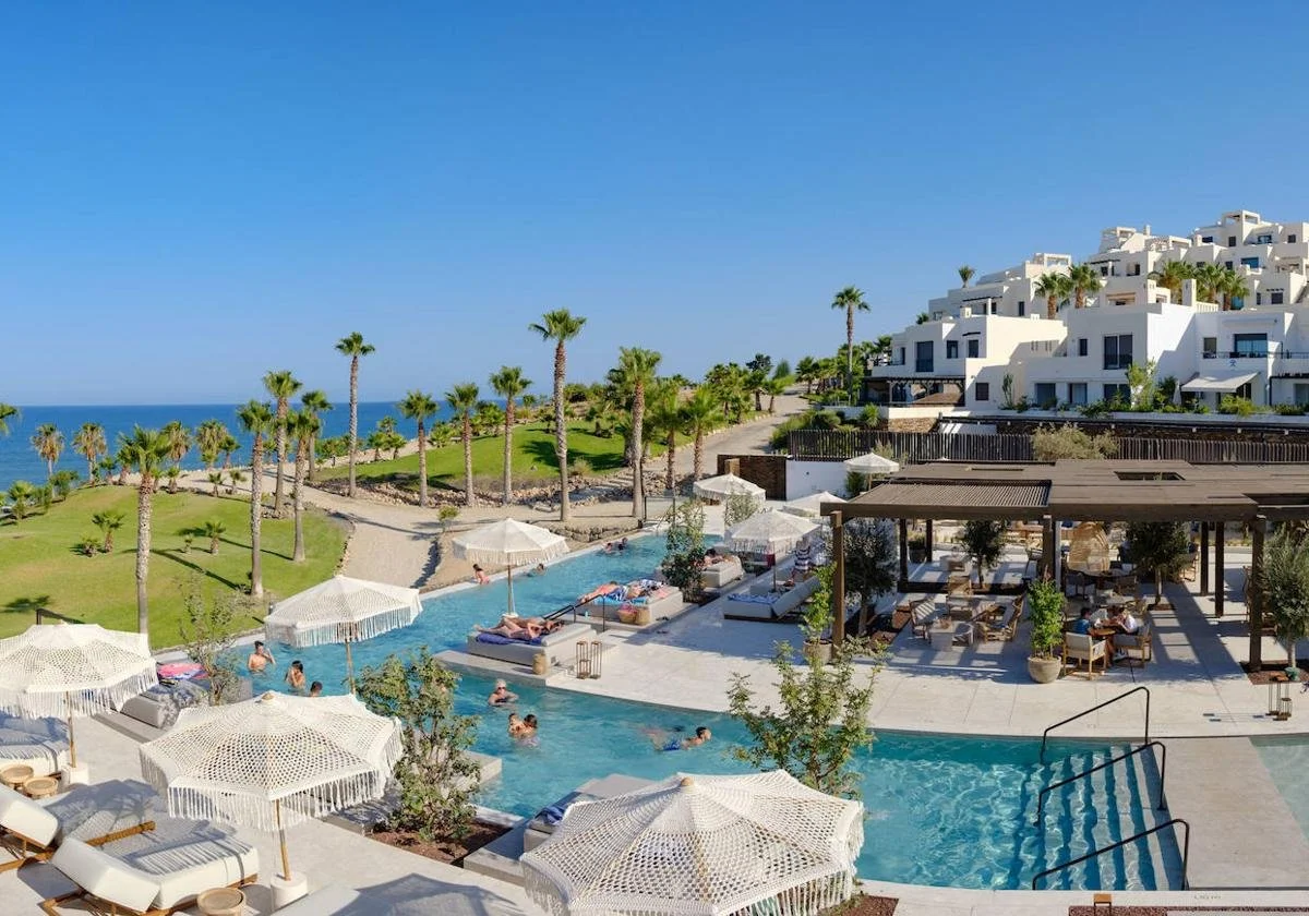 Pool area with lounge chairs and umbrellas, people swimming, lush green lawn with palm trees, modern white building complex in the background, ocean view under a clear blue sky.