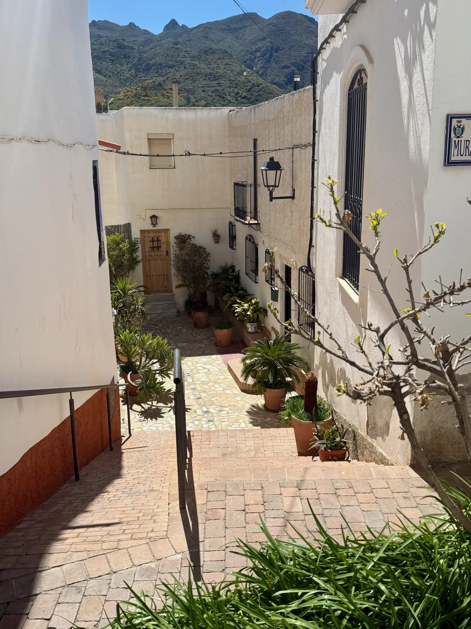 A small outdoor courtyard with potted plants along stone steps, white buildings with black window bars, a mountain range in the background, and a mountain street sign reading "MUR".