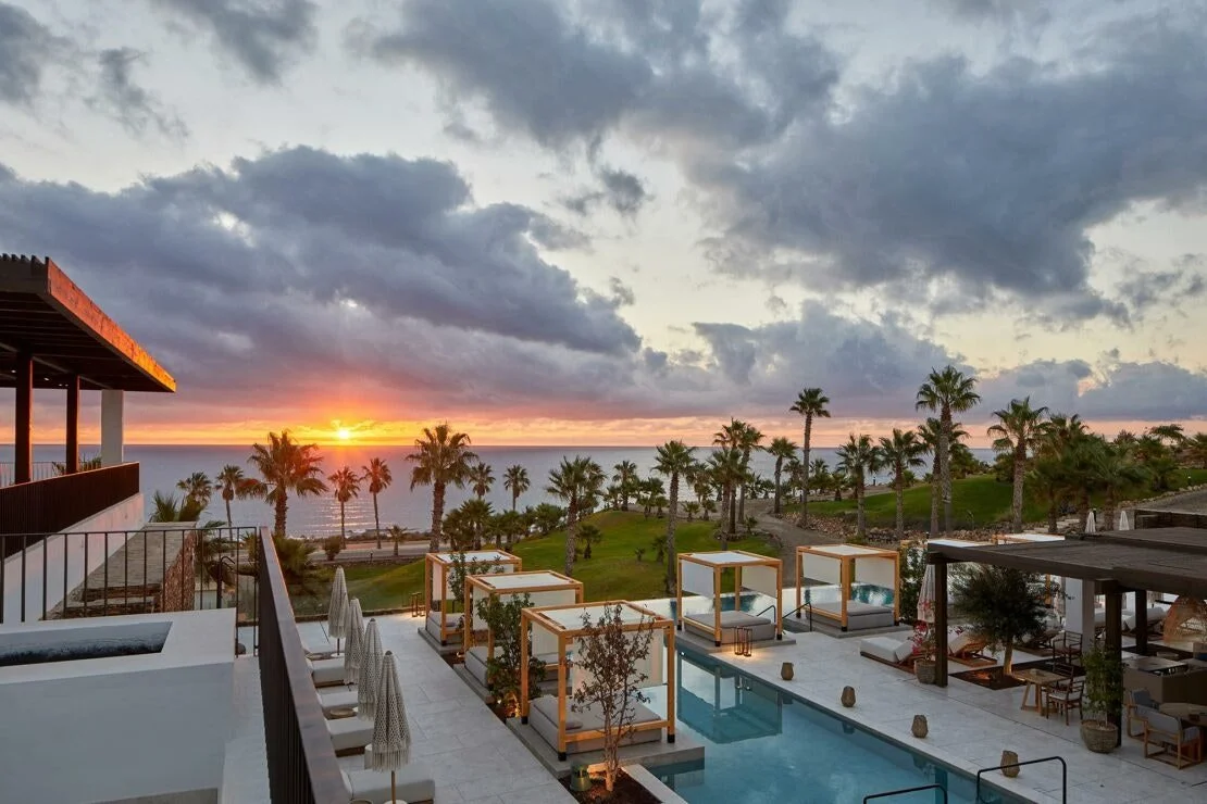 Sunset over the ocean viewed from a poolside patio with cabanas and palm trees.