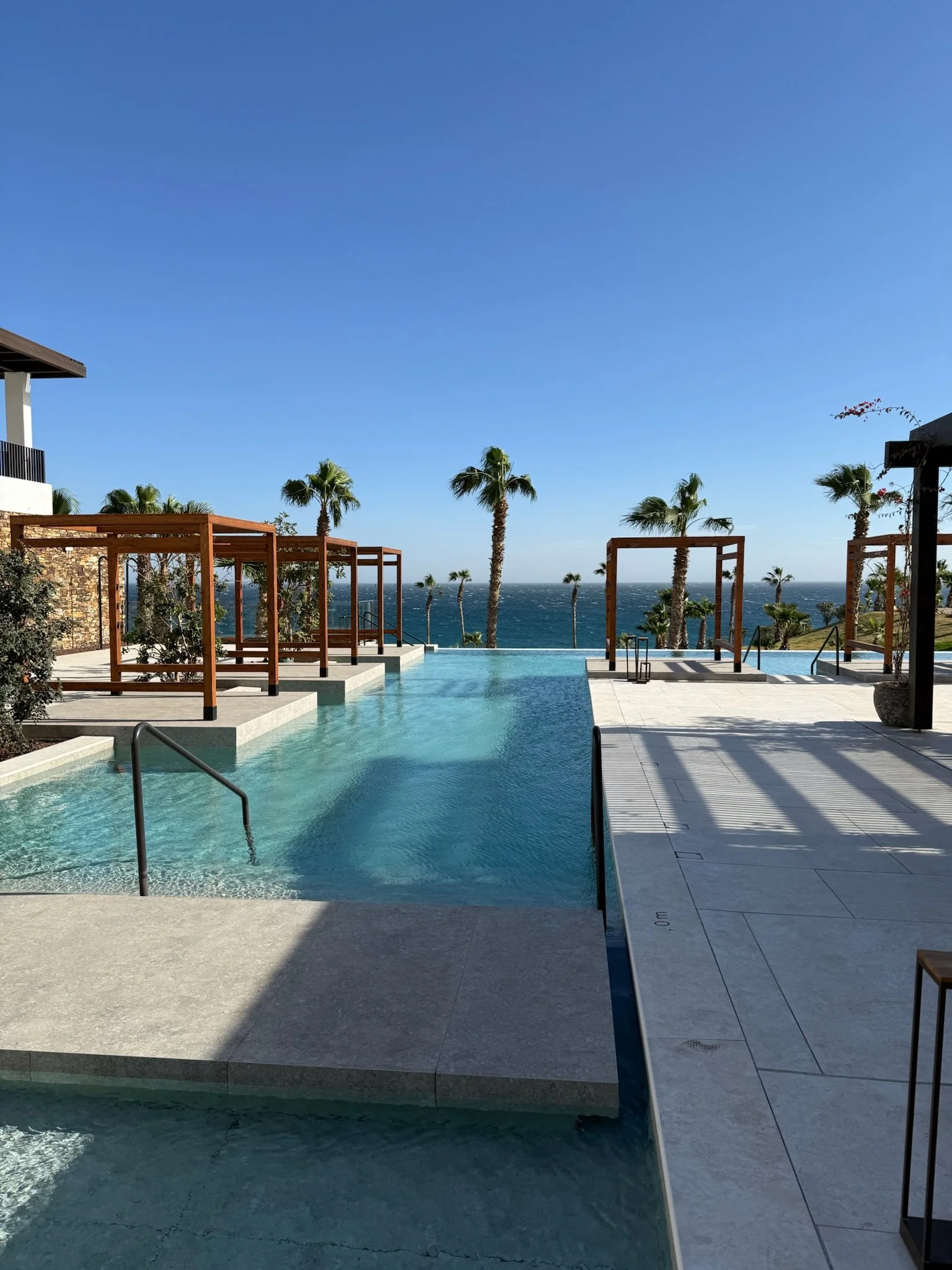 An outdoor swimming pool area with palm trees, wooden pergolas, and a view of the ocean under a clear blue sky.