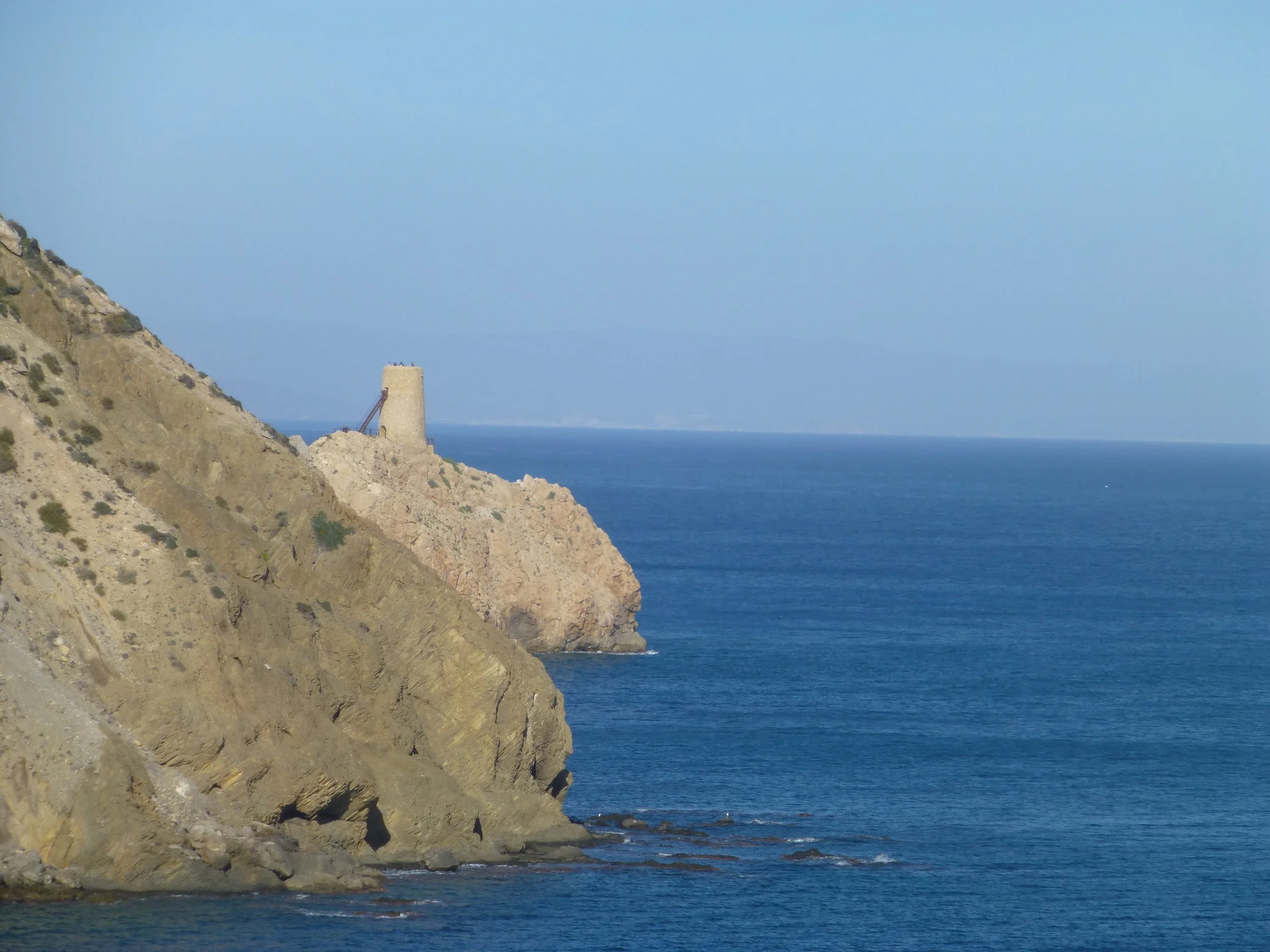 Coastal cliff with a lighthouse at the top overlooking the ocean under a clear blue sky.
