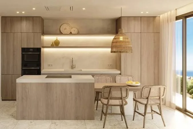 Modern kitchen with light wood cabinetry, a white island, a sink, built-in oven, and a dining table with four chairs near large windows with a view of the outdoors.