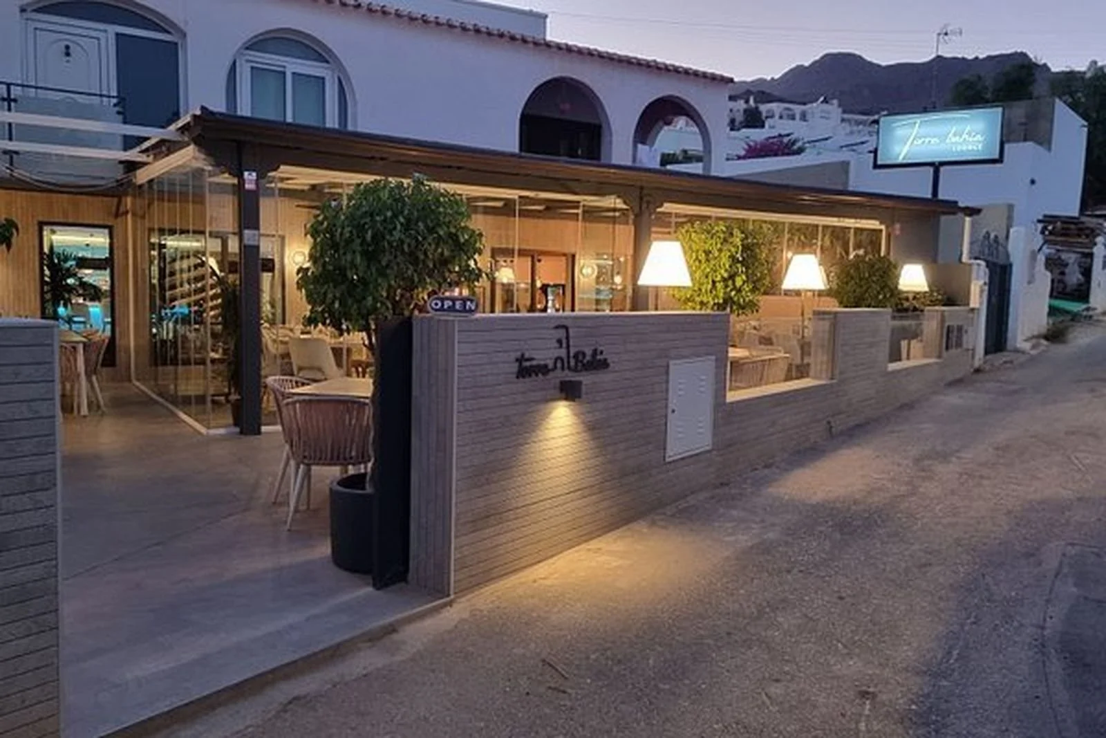 A cozy outdoor restaurant patio with warm lighting, potted plants, and a sign that reads 'Toma La Casa,' situated next to a building with arched windows and a hillside in the background at dusk.