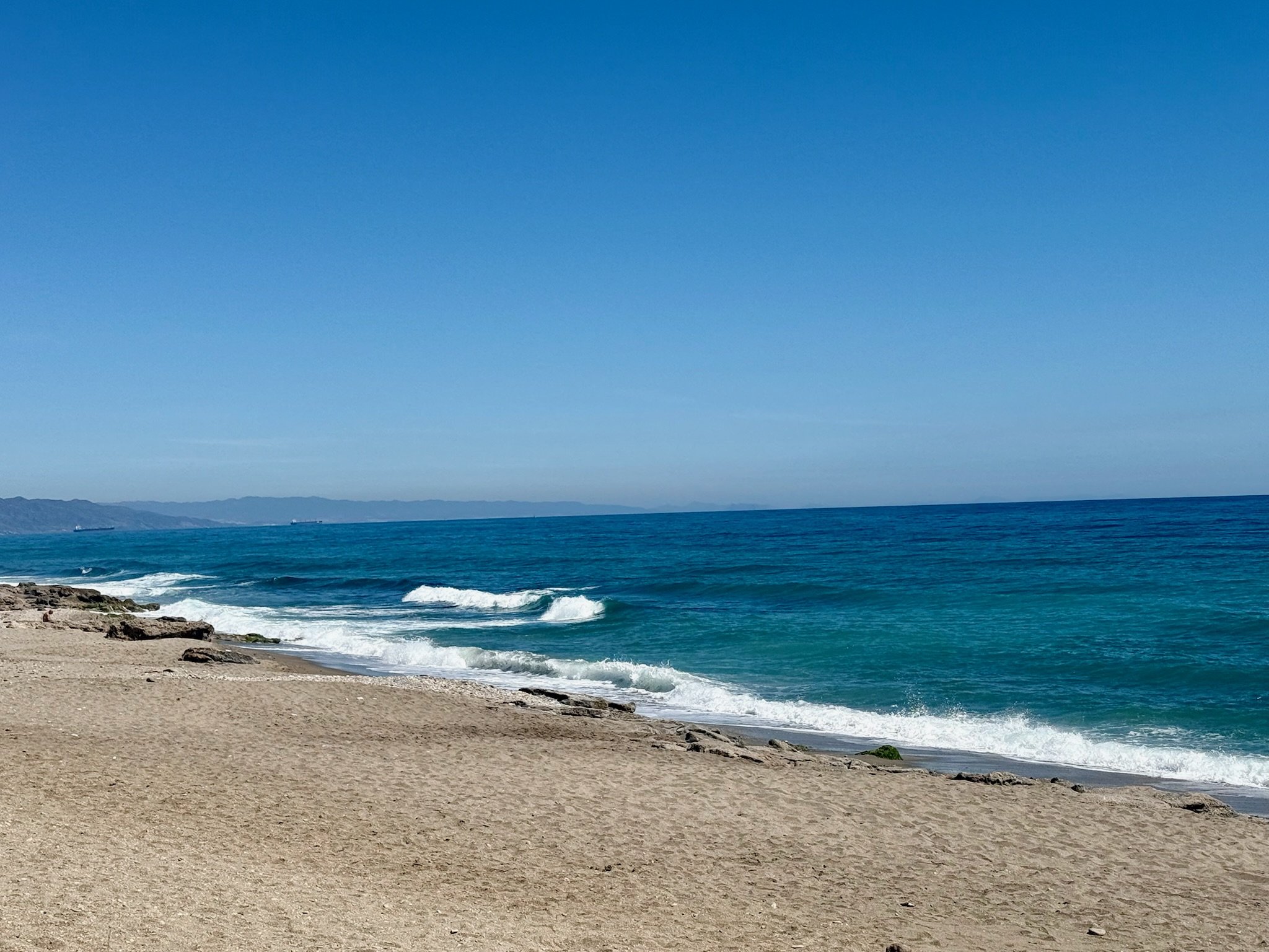 A sandy beach with small rocks, gentle waves, and a clear blue sky over the ocean with mountains in the distance.