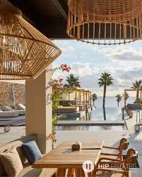 Outdoor dining area with wooden tables and chairs overlooking a pool and palm trees near the water, under thatched umbrellas.