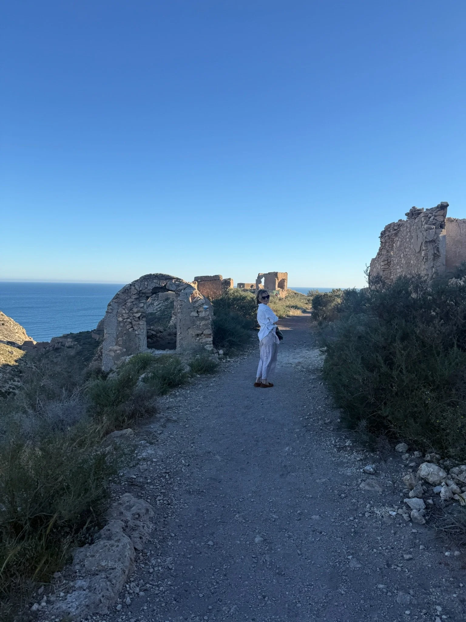 A person in a white outfit and sunglasses standing on a rocky path near ancient stone ruins overlooking the ocean under a clear blue sky.