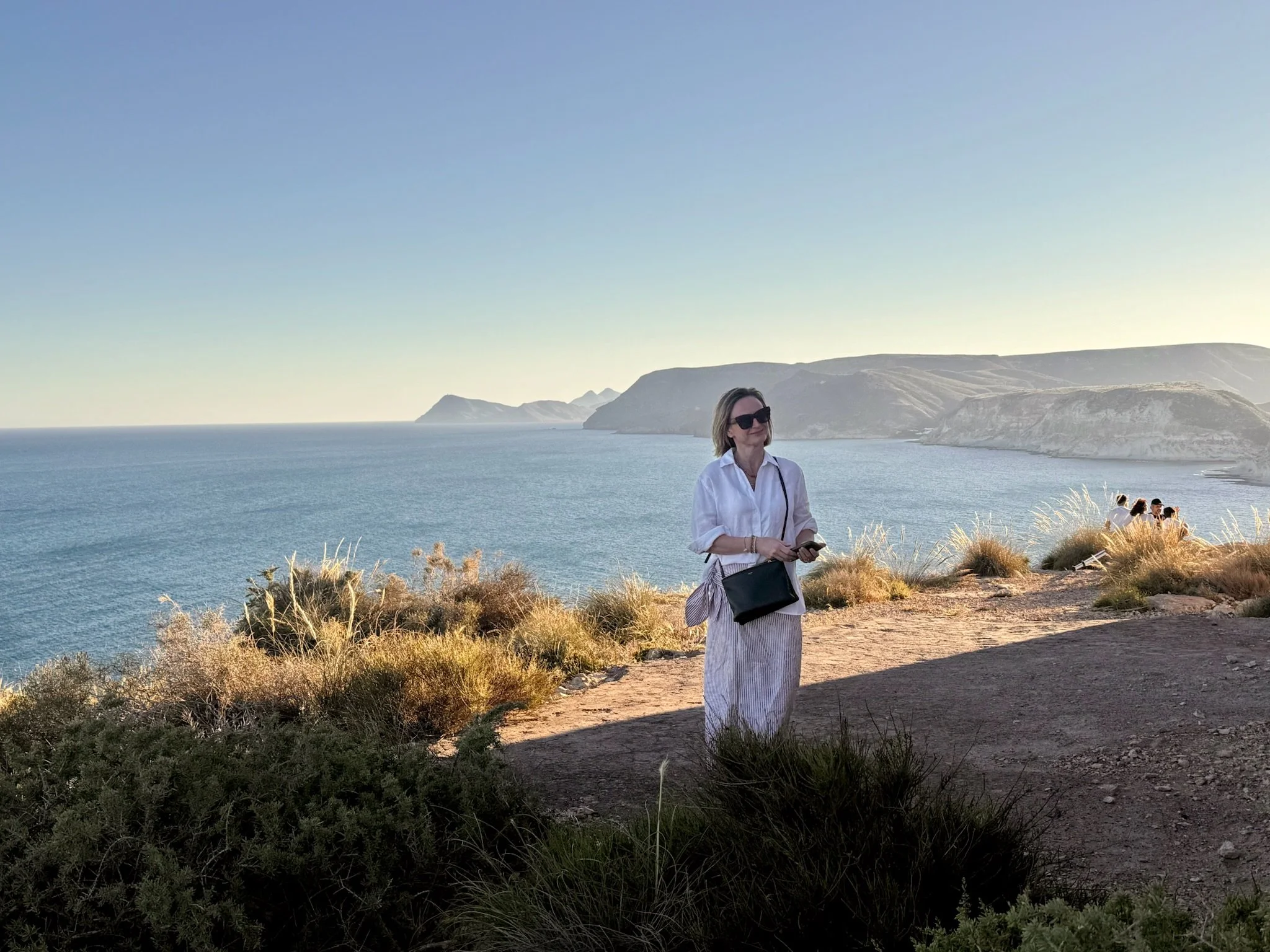 A woman with short hair and sunglasses standing by the coast with cliffs in the background, wearing a white shirt and striped skirt, holding a phone and a black bag, during sunset or late afternoon.