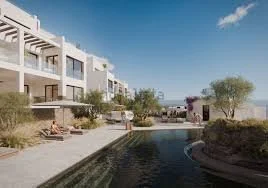 View of a modern residential complex with white buildings, a pool, and desert landscaping, under a partly cloudy sky.