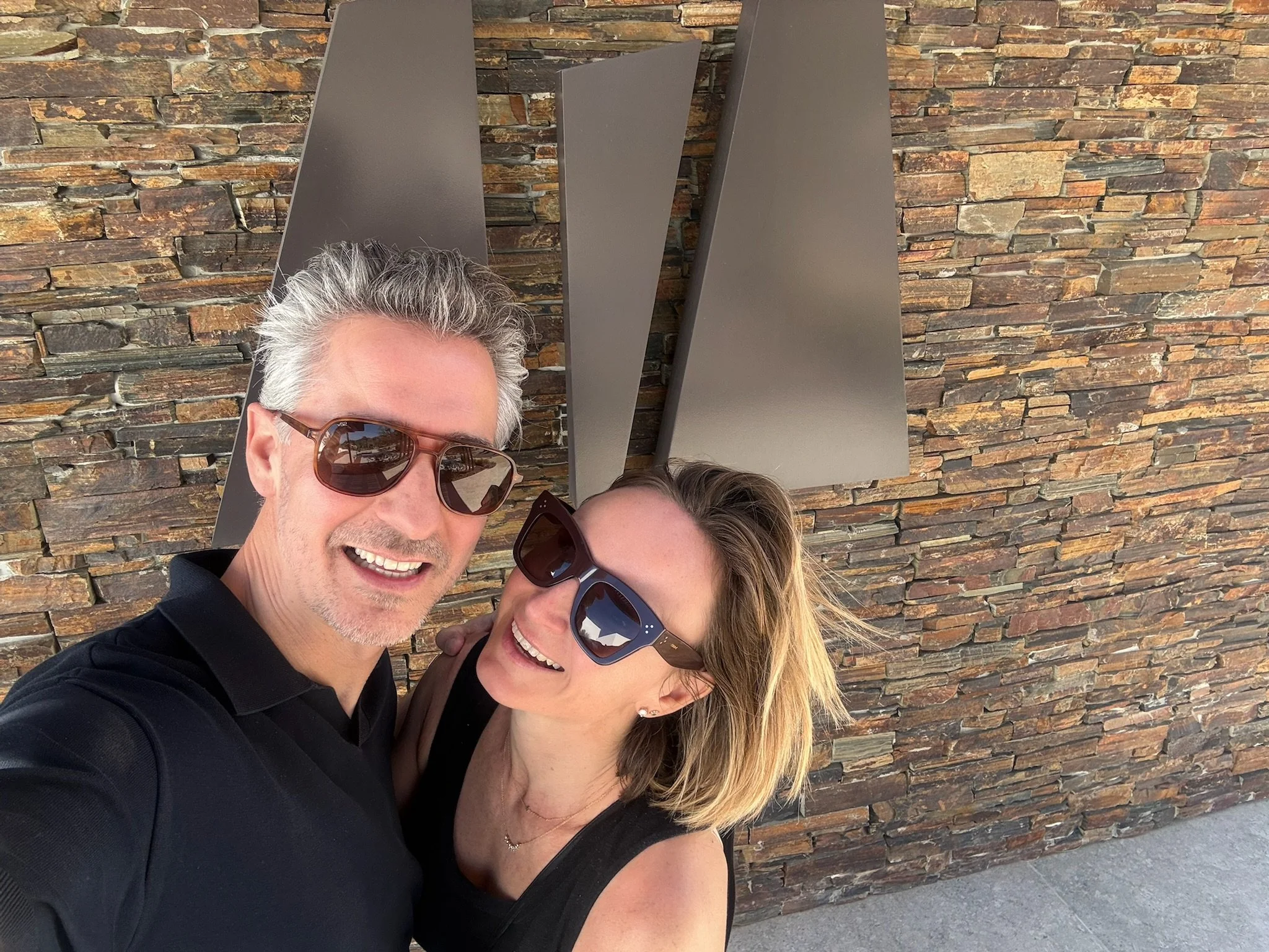 A smiling middle-aged man with gray hair and sunglasses standing next to a woman with blonde hair, both wearing black clothing and sunglasses, taking a selfie in front of a stone wall.