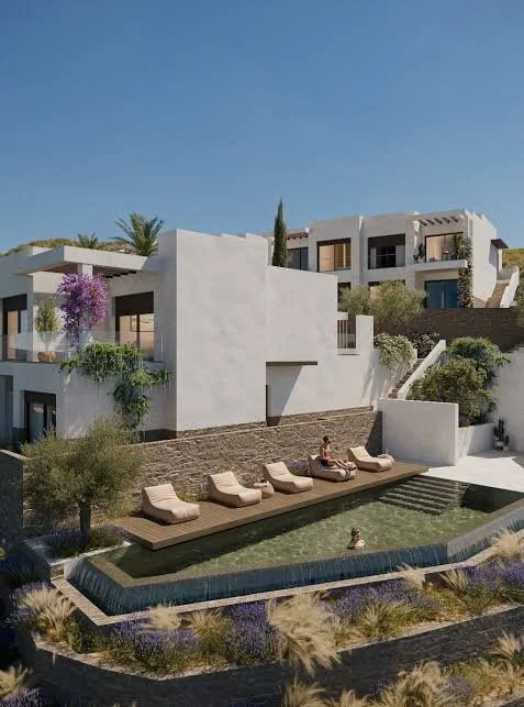 Modern white residential building with multiple levels, balconies, and terraces, surrounded by greenery and a pool area with lounge chairs and water feature.