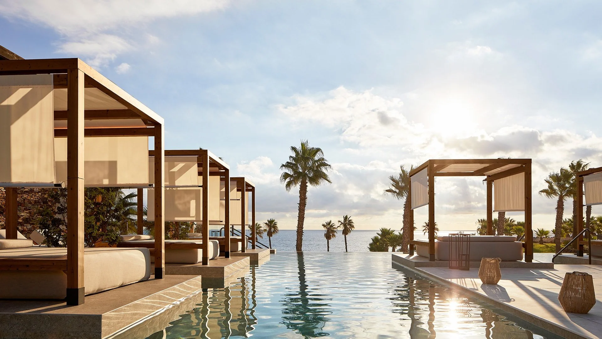 Luxury resort pool area with modern cabanas, palm trees, and ocean view in the background during sunset.