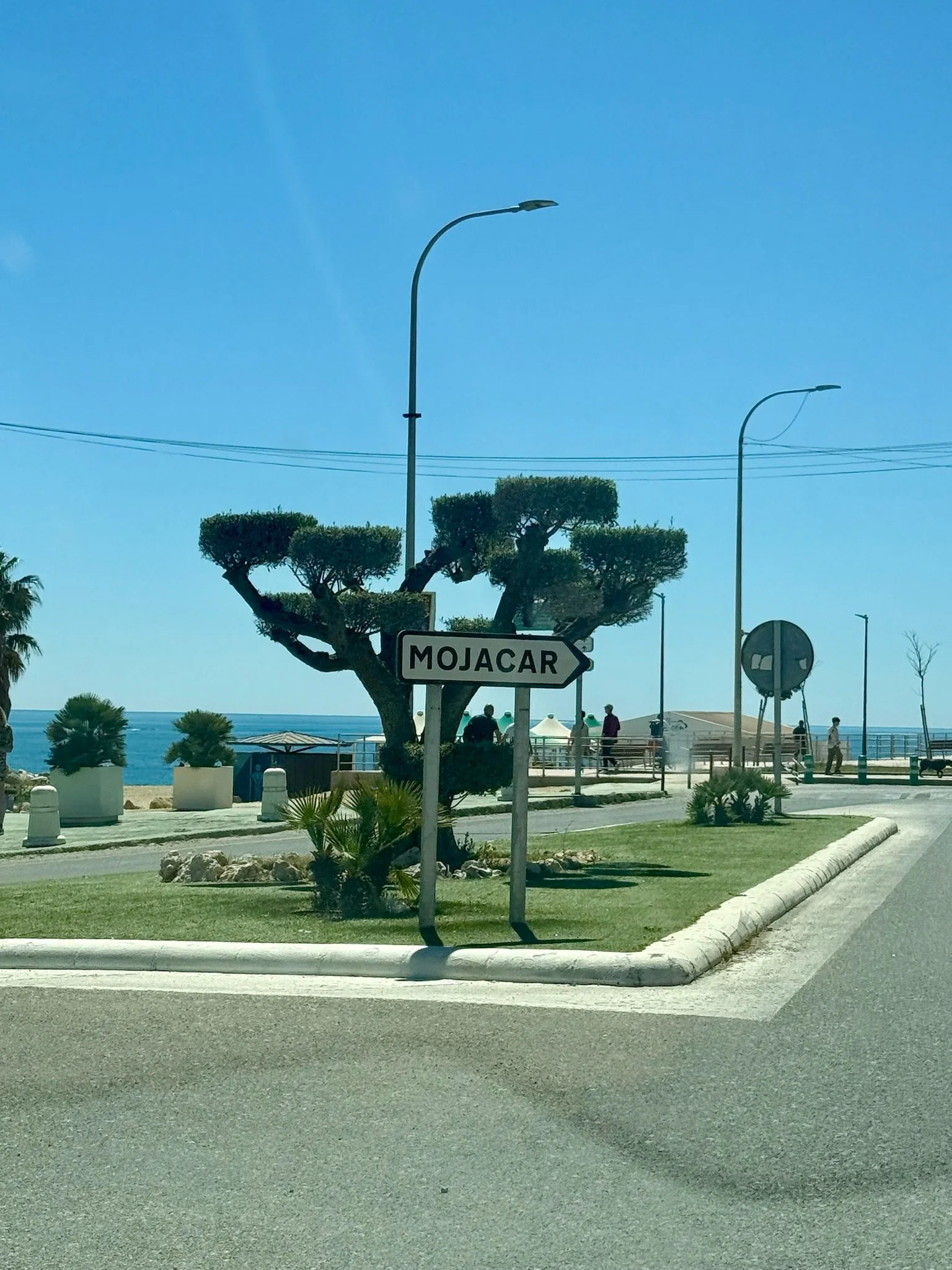 Street scene near a beach with a sign pointing to Mojacar, ocean view in the background, and people walking along the promenade.
