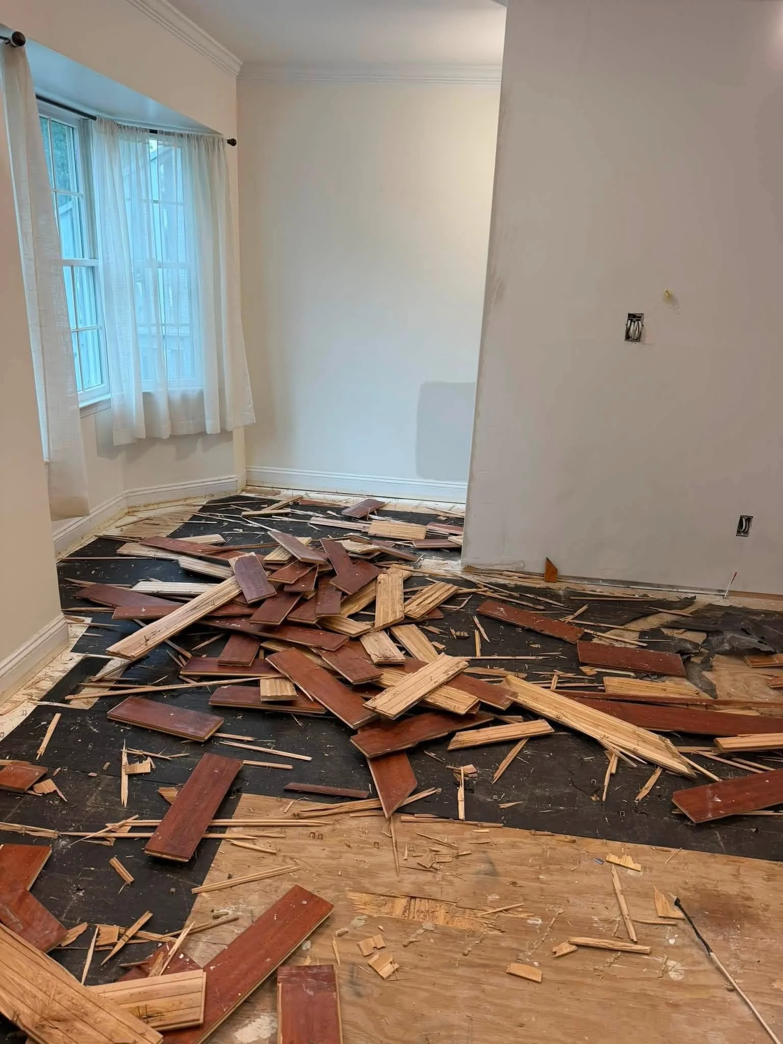 A room with hardwood floors and a window with white curtains. The wall to the right shows signs of damage with a hole and exposed wires. The floor is covered in broken wooden pieces, likely from flooring or furniture.