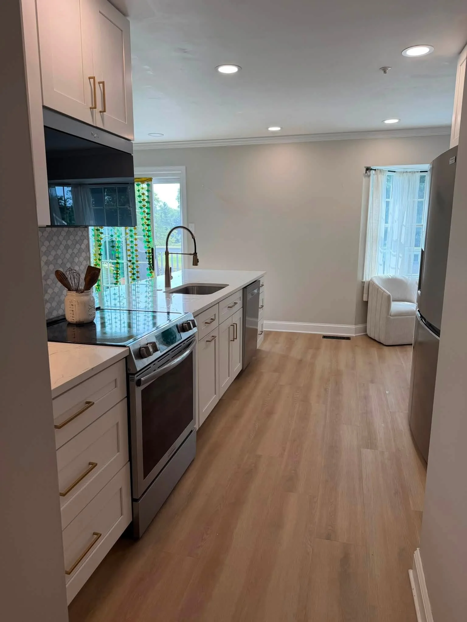 Modern kitchen with white cabinets, stainless steel appliances, a white marble countertop, a black stovetop, a sink with a high-arch faucet, a window with white curtains, a small beige armchair, and hardwood flooring.