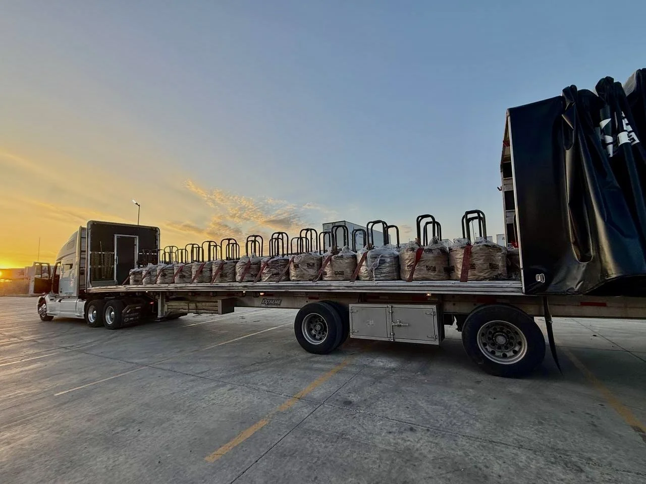A semi-truck parked in an open lot during sunset, carrying bags of supplies with hand trucks on the flatbed.