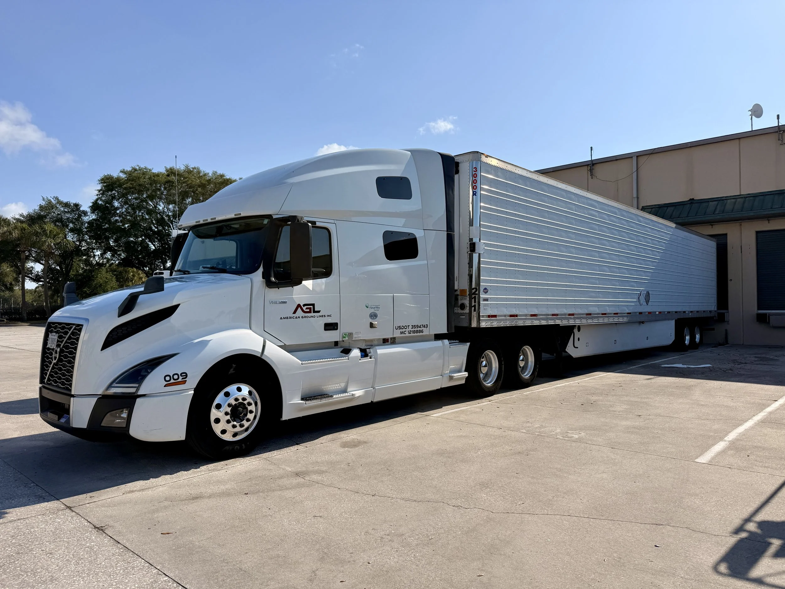 A large white semi-truck parked in an empty parking lot outside a beige building with a green roof on a clear day.