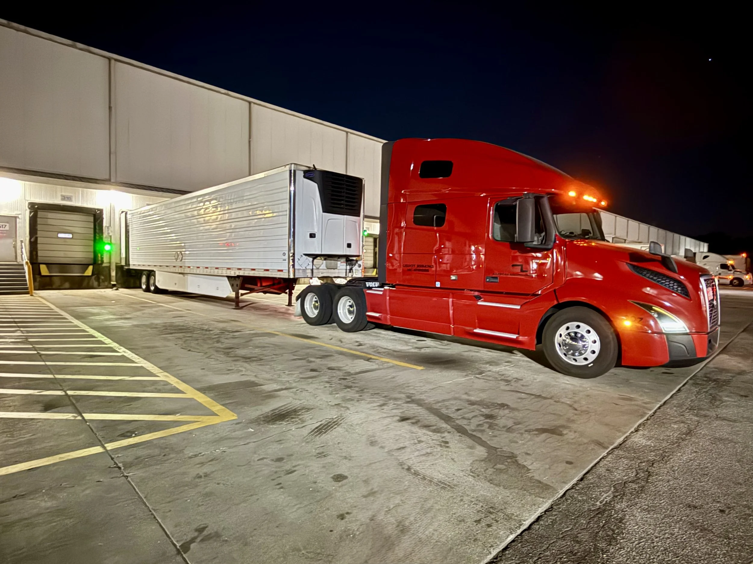 A red semi-truck parked outside a warehouse at night, with a white trailer attached, next to loading docks with closed doors.