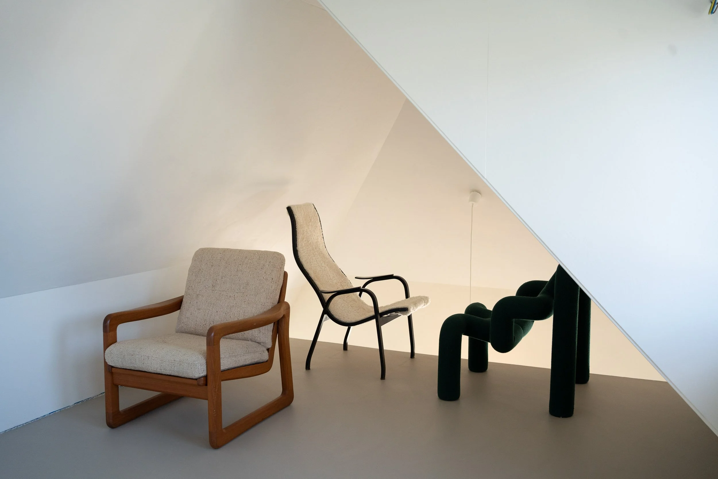 Drie verschillende stoelen in een minimalistische kamer met een witte muur en een plafond met schuine hoek.