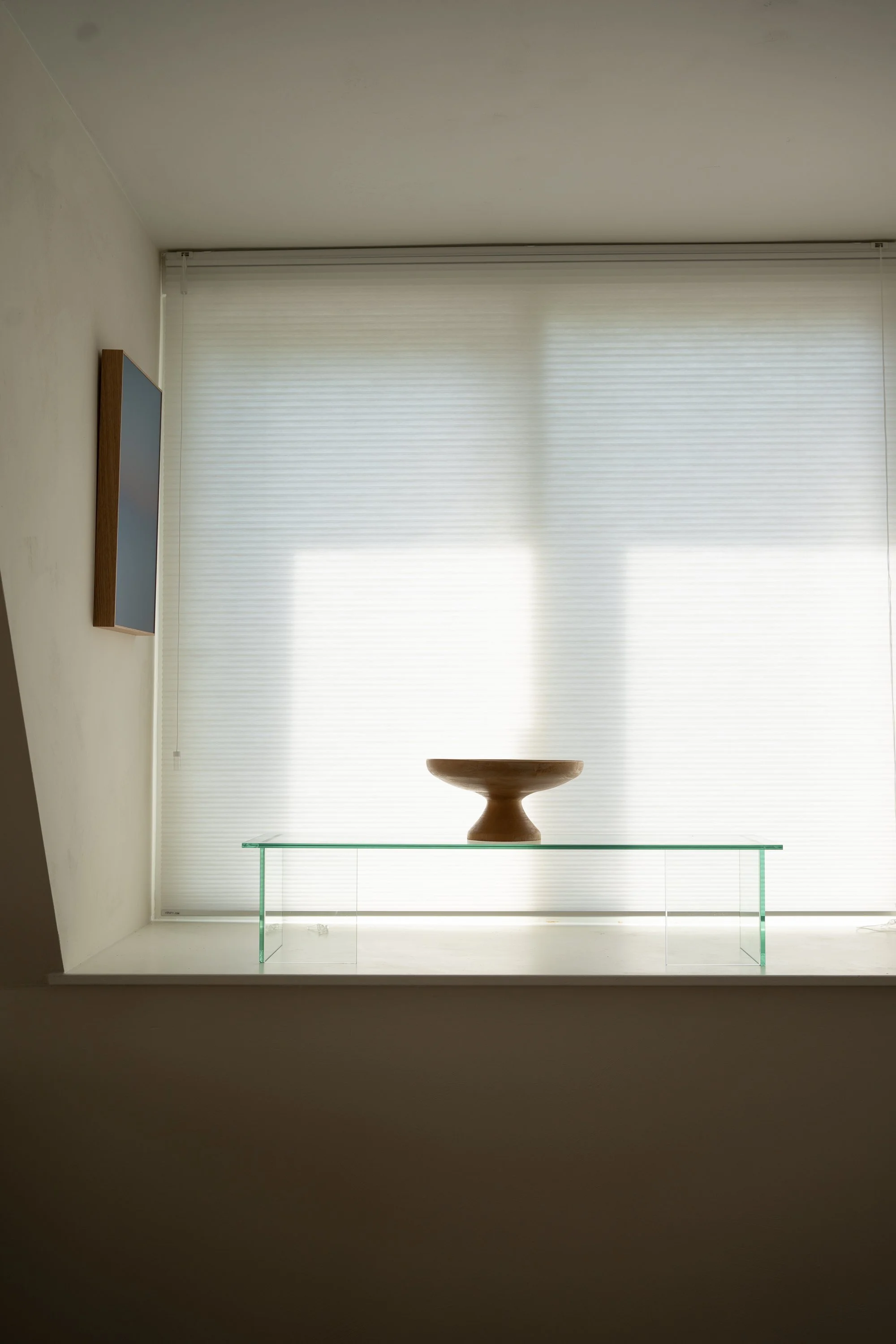 Een witte vensterbank met een glazen tafel en een houten kommetje, tegen een raam met witte jaloezieën.