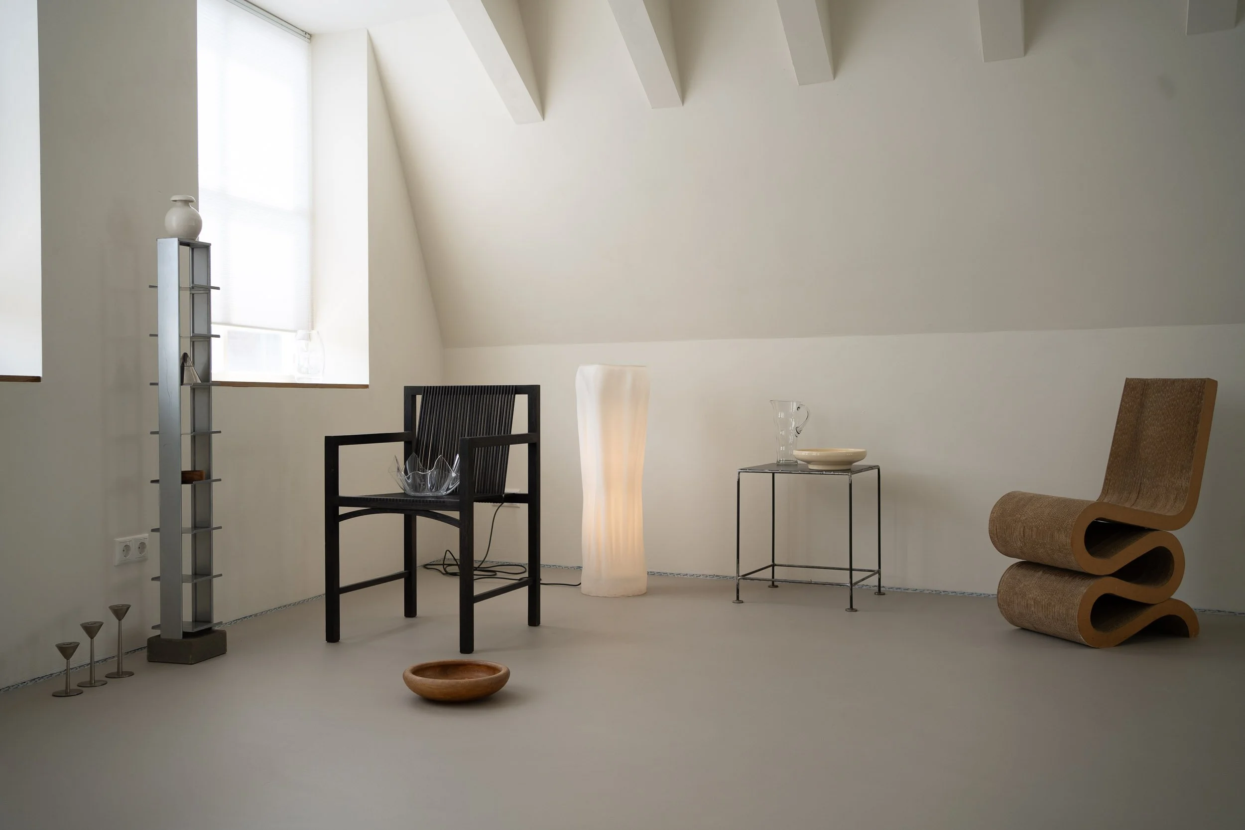 Interieur van een minimalistisch huis met witte muren en meubels, waaronder een zwarte stoel, een houten tafeltje, en verschillende decoratieve objecten zoals vazen, een tafel met glas en keramiek, en een uniek wervelvormig bankje.