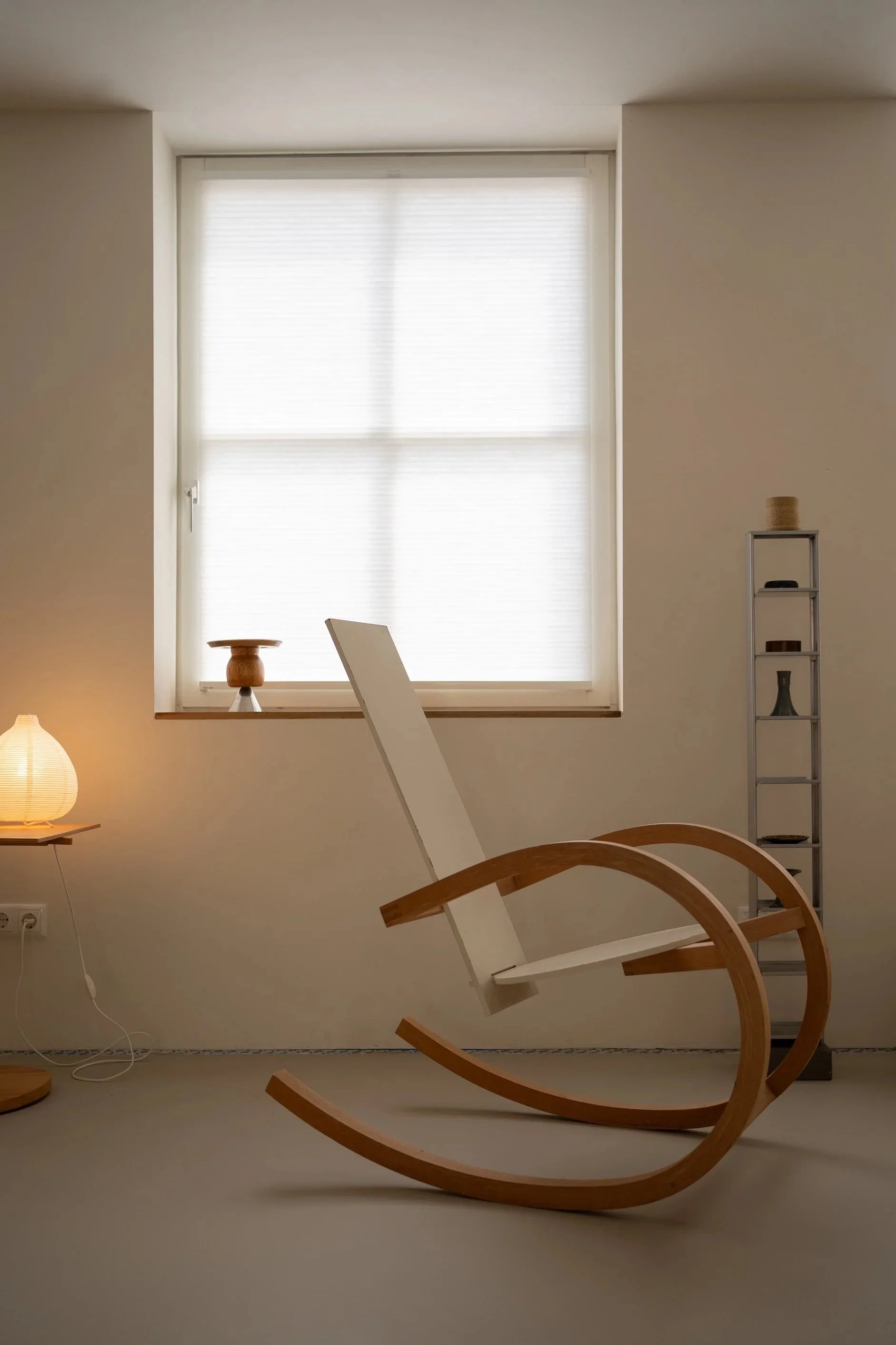 Een kamer met een grote witgelakte houten stoel die op z'n kop ligt, een groot raam met een witte raamtrek en een rolgordijn, een wit rek met decoraties, een kleine houten tafel met een lamp, en een stroomkast met stekkerdoos.