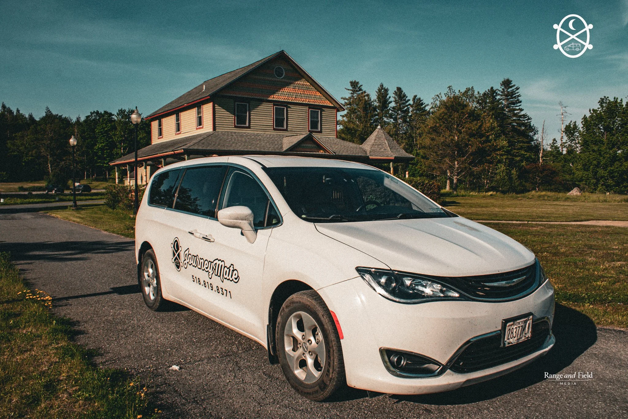 A white van with 'JourneyMate' logo and contact information parked on grass beside a paved area in front of a large, historic house with a steep roof and multiple windows, surrounded by trees and lamp posts.