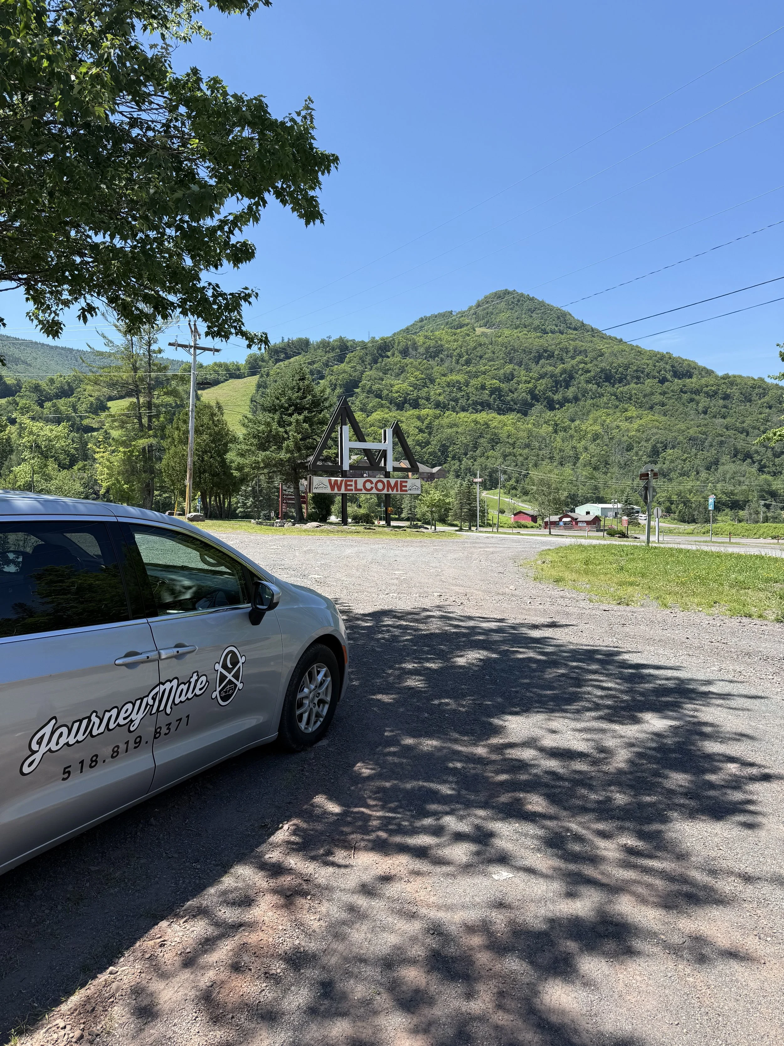 A scenic view of a mountainous area with a clear blue sky, green trees, and a small town with buildings and a welcome sign in the background. There is a parked car in the foreground, and shadows from the trees cast onto the dirt ground.