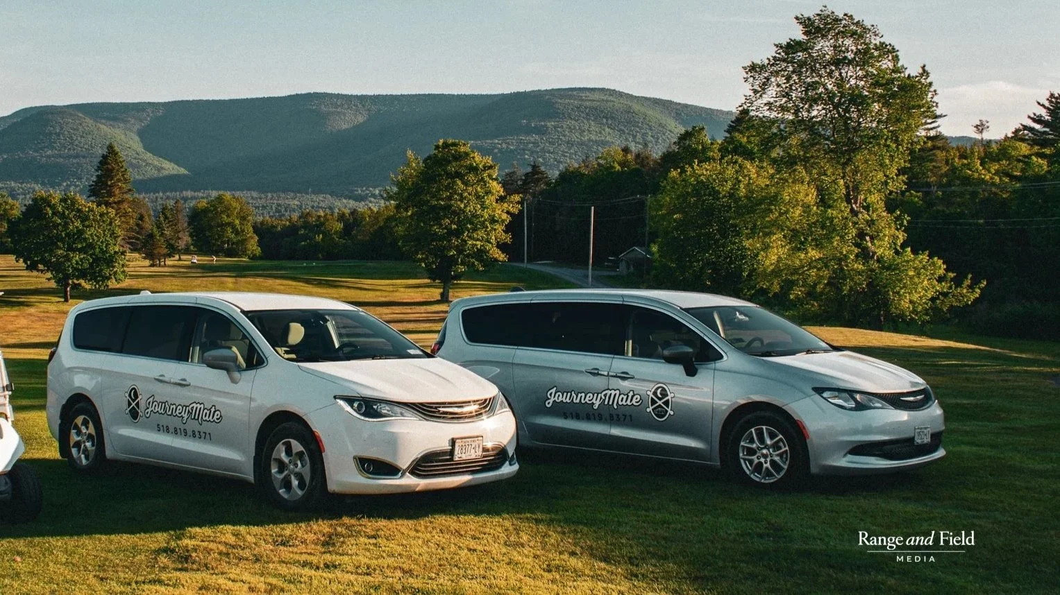 Two white vans parked on a grassy hill with trees and mountains in the background. The vans have 'JourneyMate' logos and phone numbers on their sides.