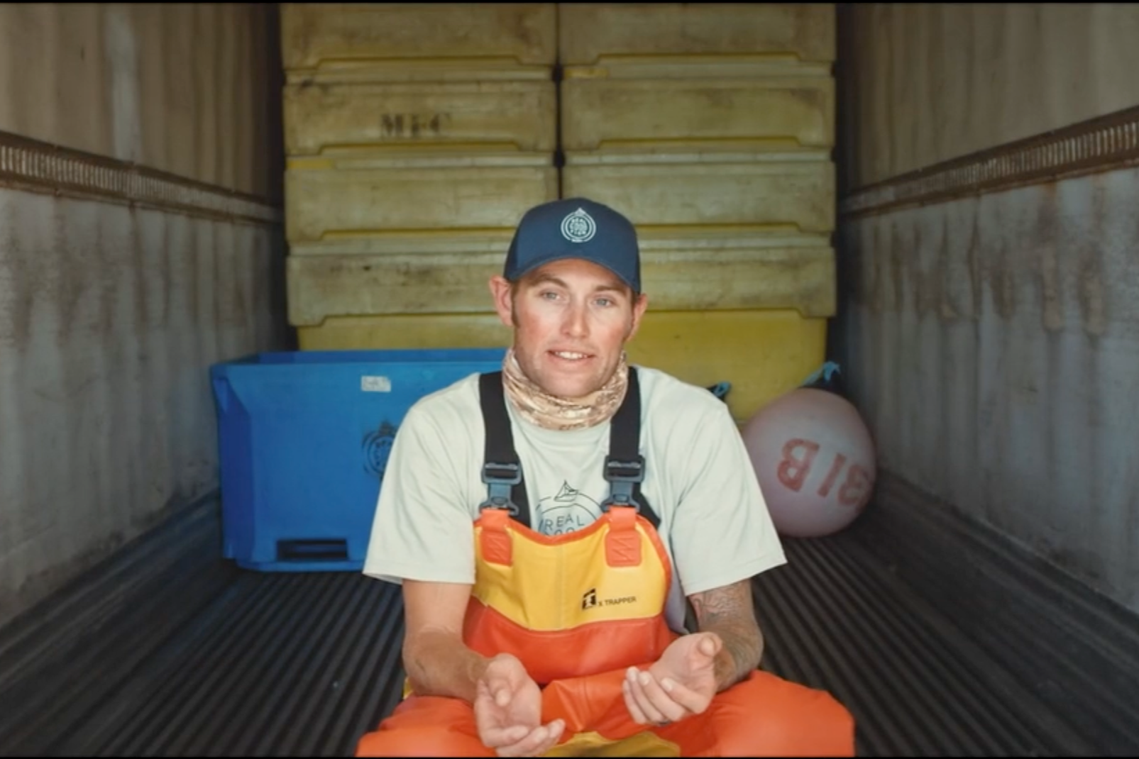 A man sitting inside a shipping container, wearing an orange life jacket, a blue cap, and a beige shirt, with blue containers and a ball labeled 'BIB' inside the container.