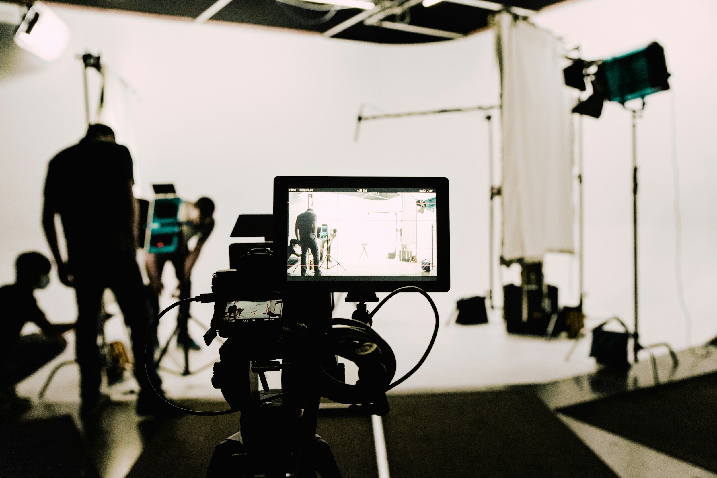Studio setup with camera on tripod filming a person standing in front of a white backdrop, surrounded by lighting equipment and crew members.