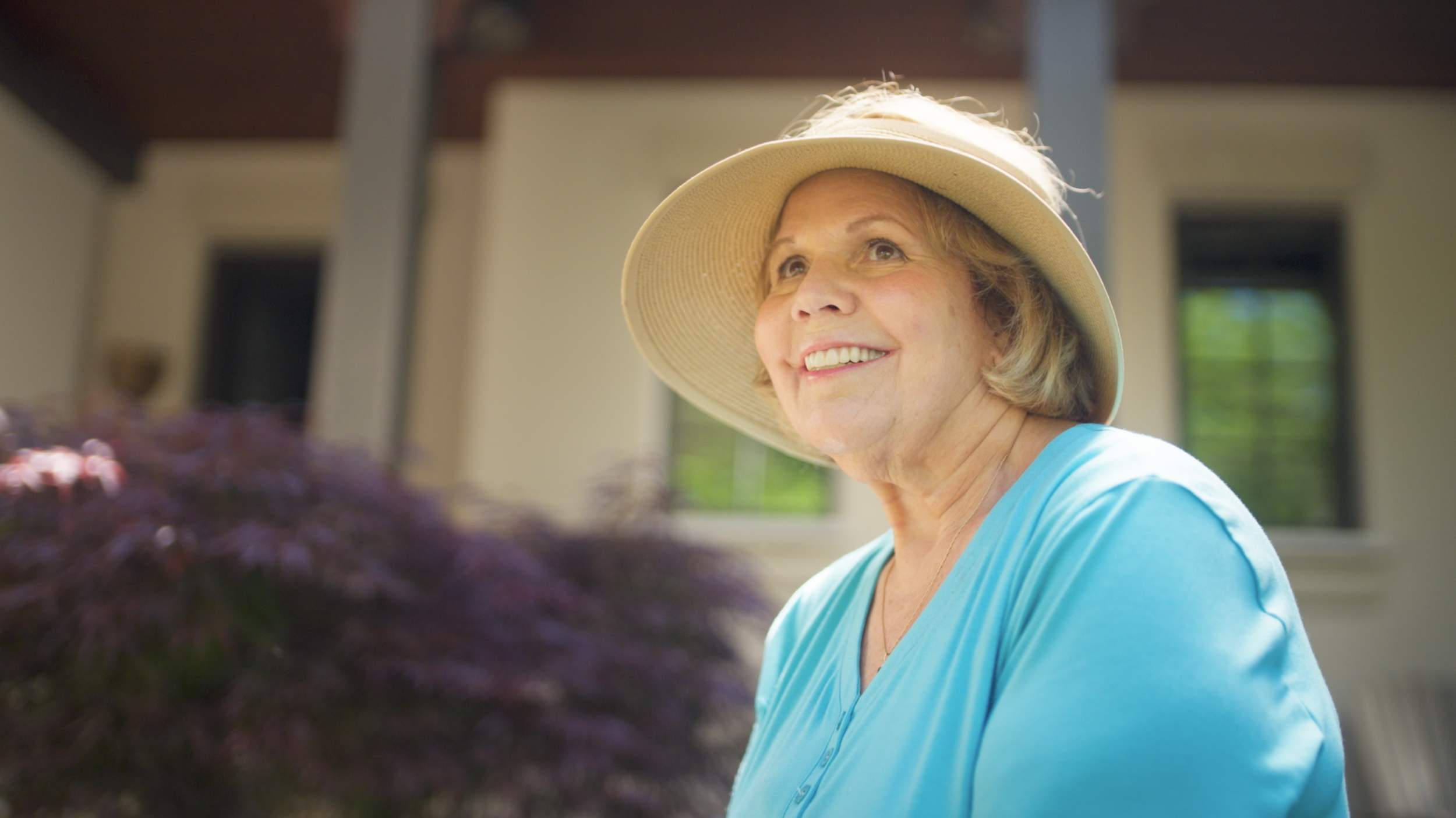 Smiling elderly woman with gray hair wearing a wide-brimmed sun hat and light blue shirt indoors, with sunlight streaming through windows in the background.