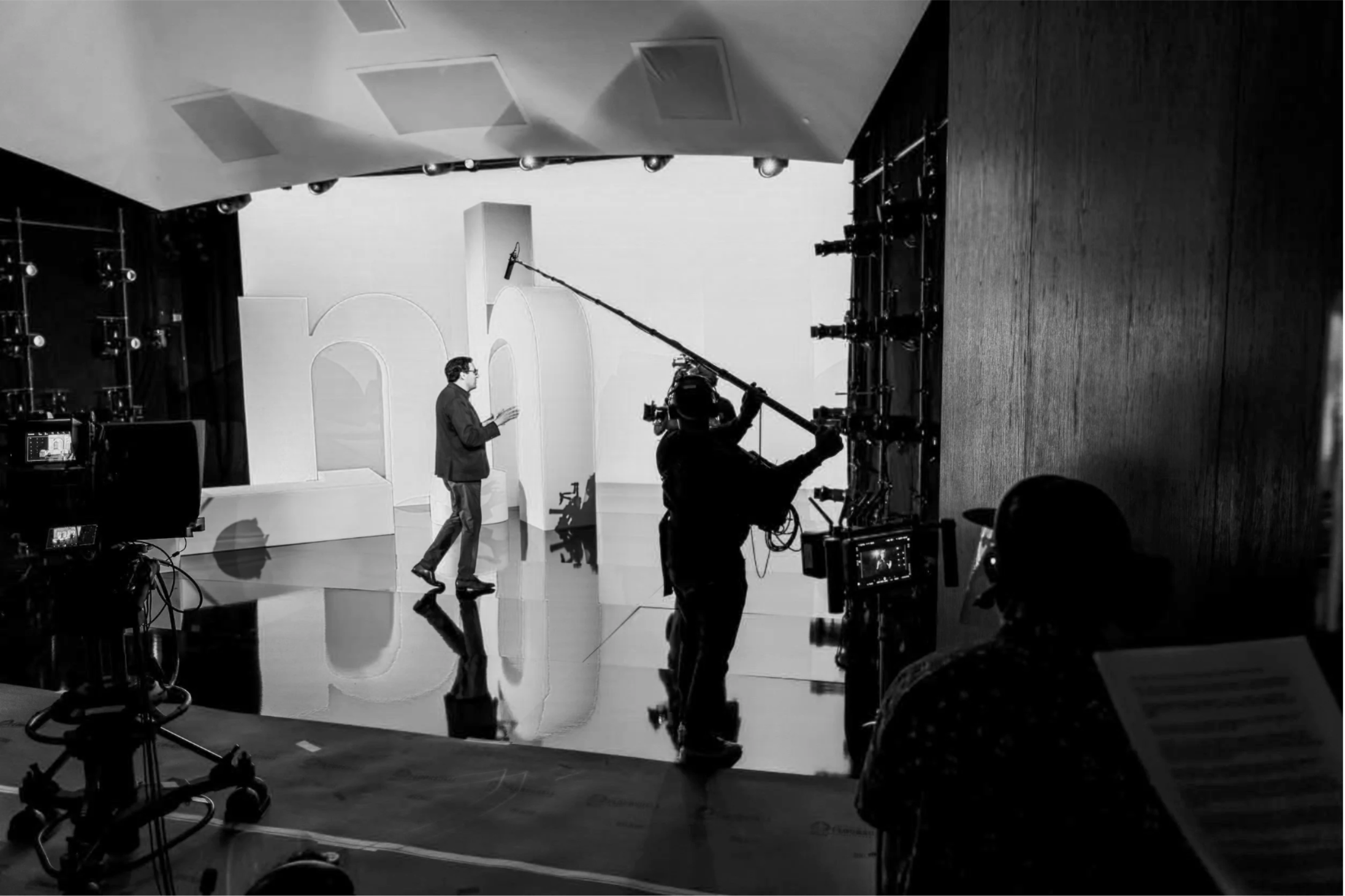 A man in a suit standing on a television studio set with a white background and large numbers "50" in the background. A camera operator is filming him while a boom microphone hangs overhead. Equipment and lighting are visible around the set.